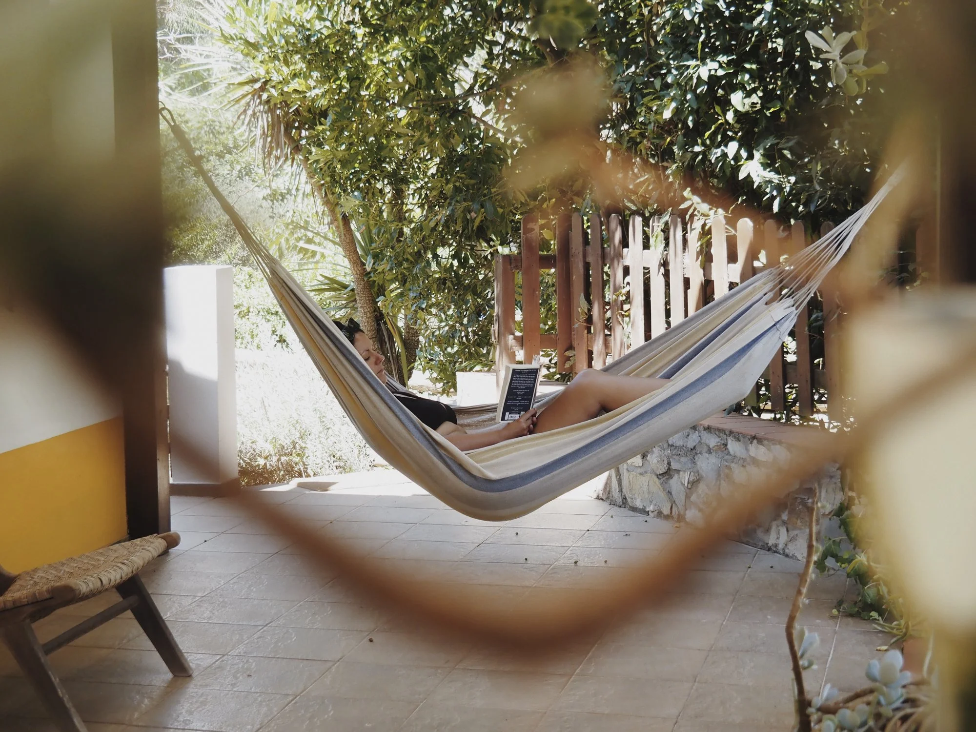 Person relaxing on a hammock in an outdoor setting with lush green trees and a wooden fence in the background.