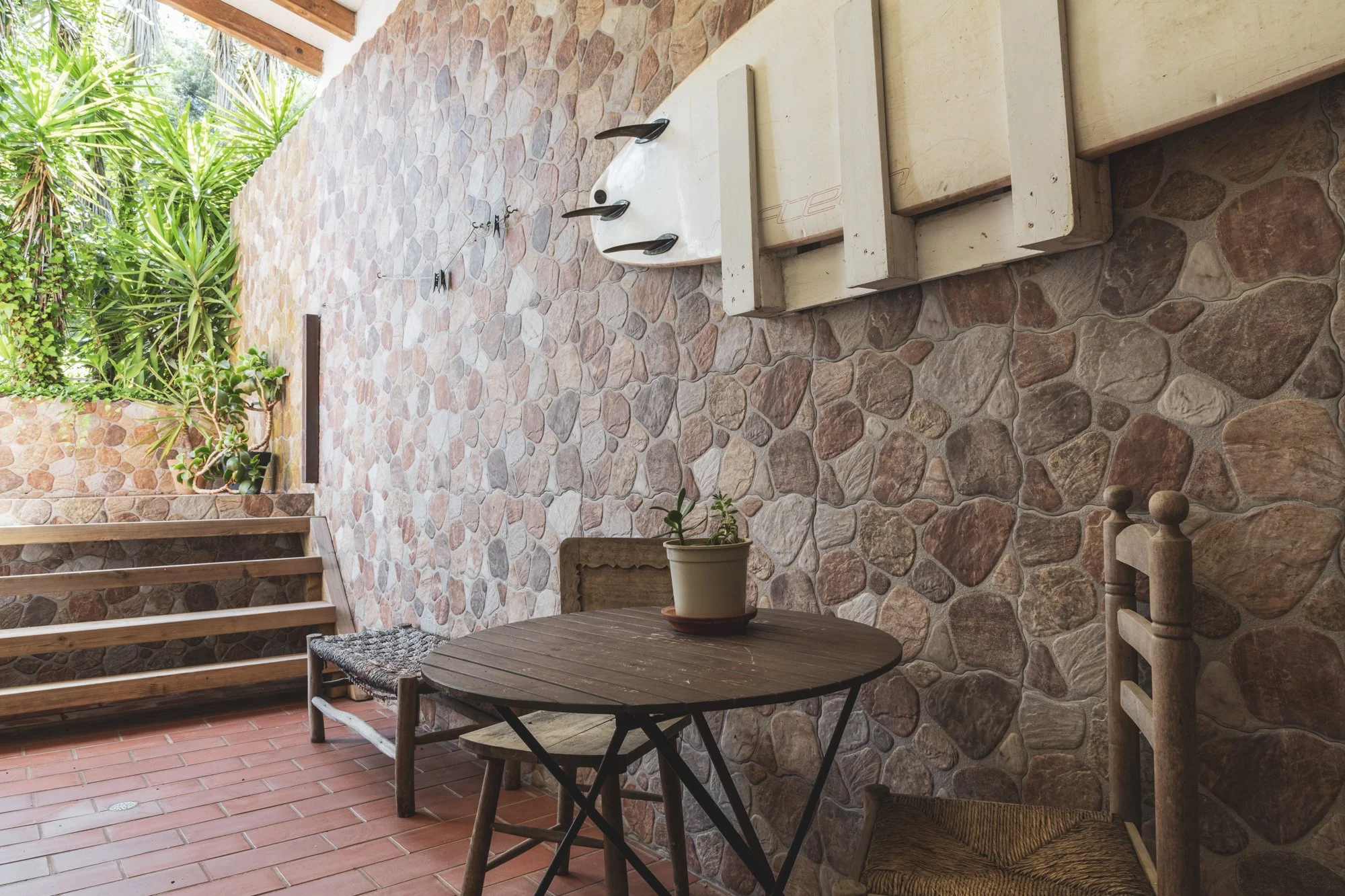 A covered patio area with a stone wall and tiled floor. There is a small wooden table with a potted plant, a wooden bench with a woven seat, and stairs leading up to another level. Lush green plants are visible outside.