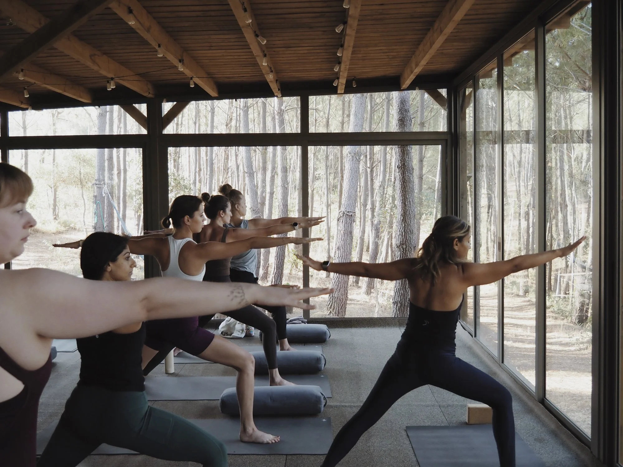Group of women participating in yoga class in Portugal retreat near large windows with a view of trees outside, following the instructor's pose.
