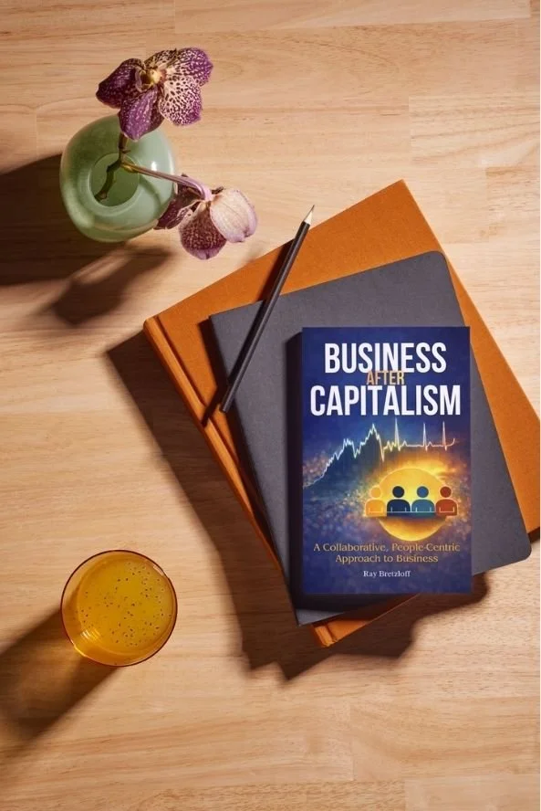 A desk with a book titled 'Business After Capitalism', a black notebook, an orange notebook, a pen, a glass of orange juice, and a small vase with purple orchids.