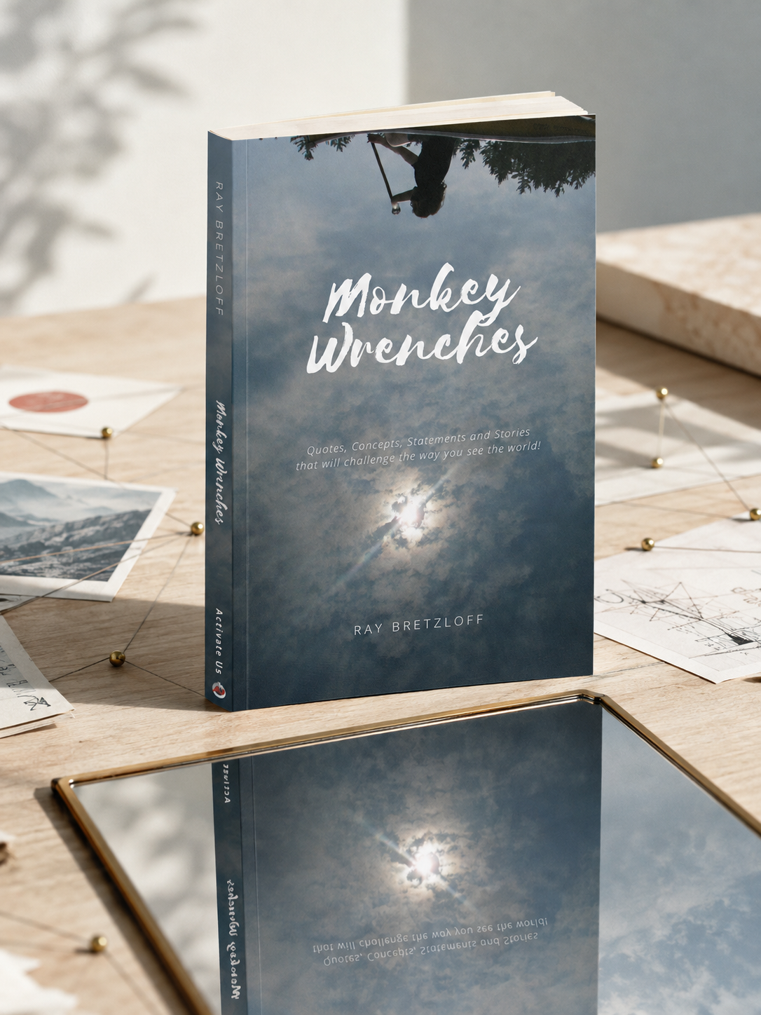 A book titled 'Monkey Wrenches' by Ray Bretzloff is standing upright on a wooden table, with its cover showing a sky with clouds and sunlight. There are various papers and photographs scattered on the table around the book, and a mirror reflecting the sky and part of the book.