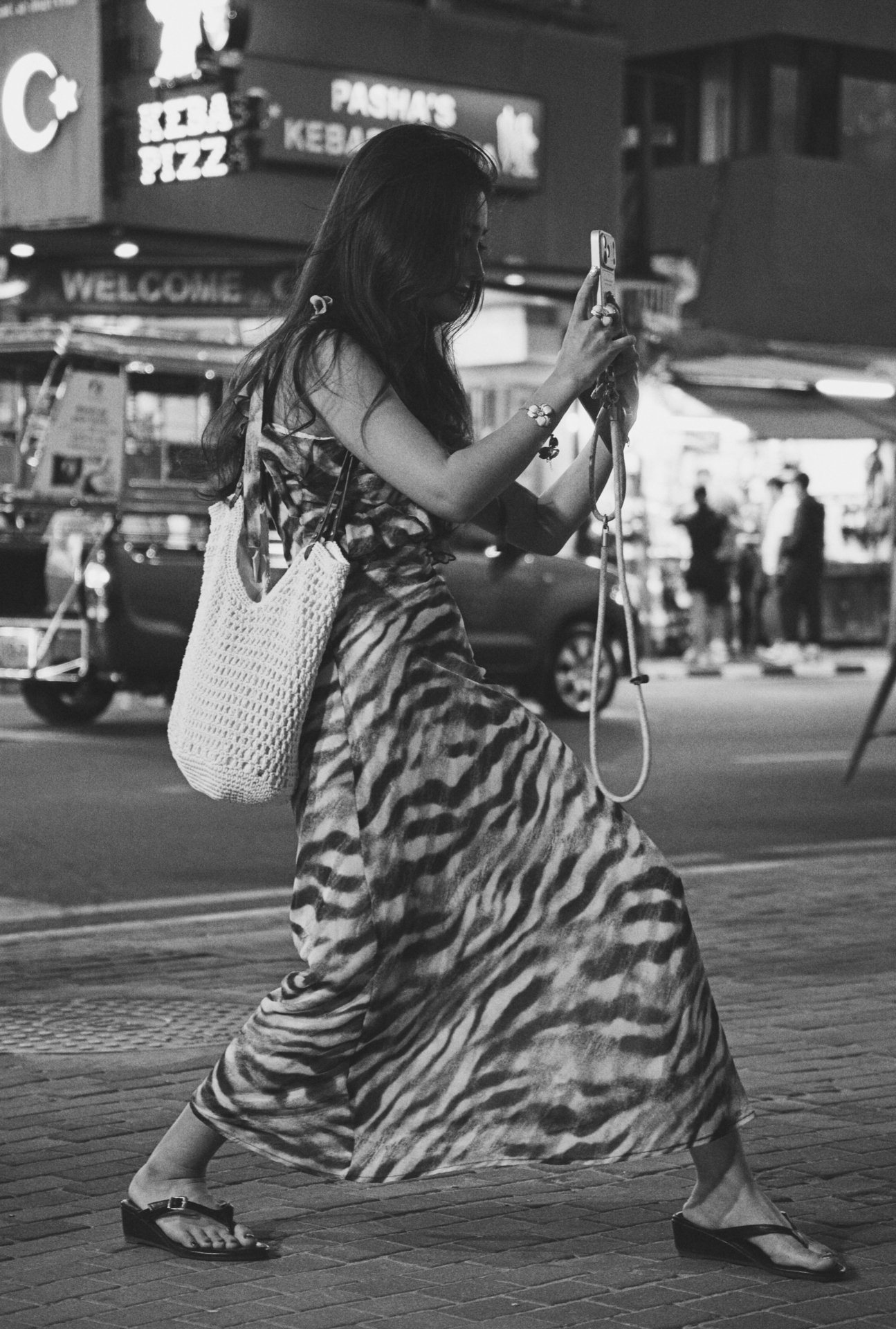 A woman with long dark hair wearing a zebra stripe dress and sandals is crossing a street at night, using her phone. She has a woven bag hanging on her shoulder. There are signs and shops in the background.