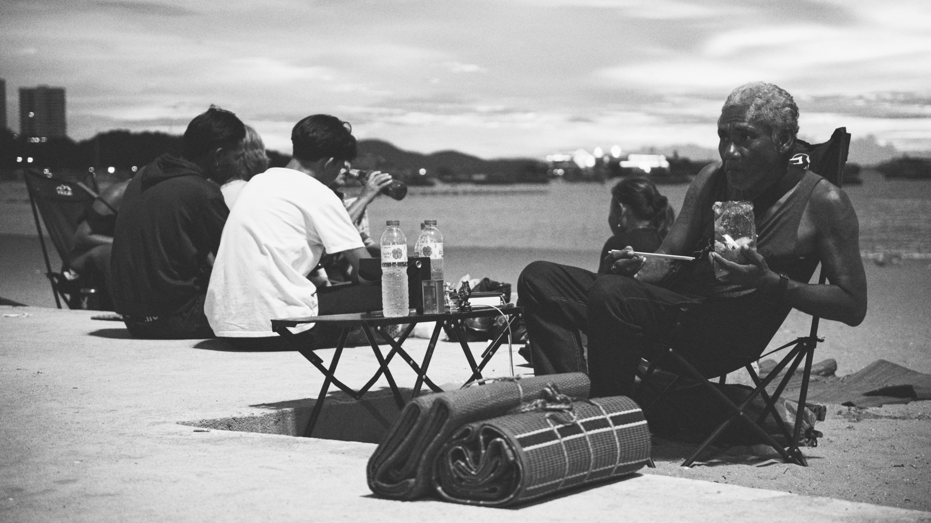 A group of people sitting outdoors on folding chairs and a table, some drinking water or soda, during evening or sunset, with mountains and city lights in the background; black and white photo.