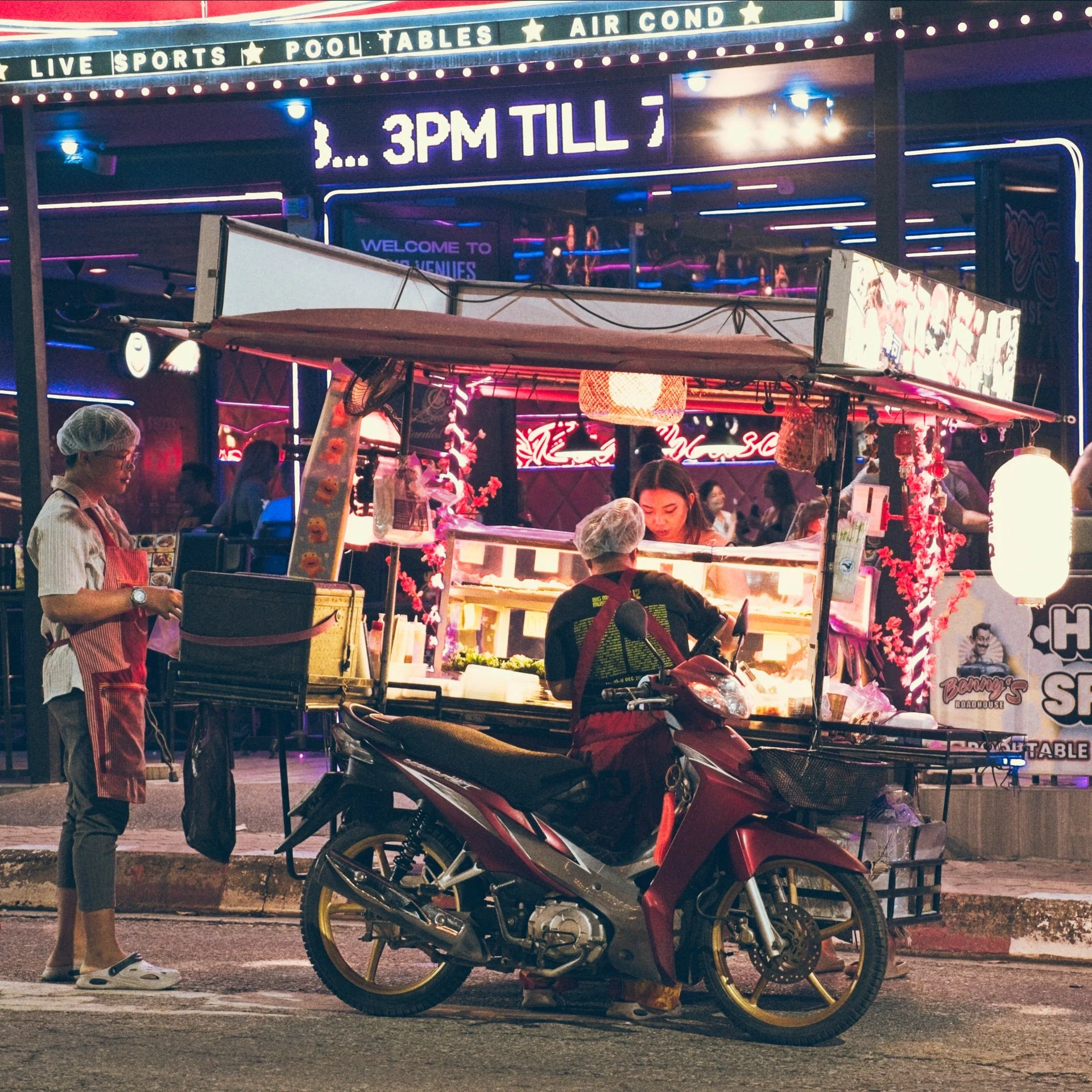 A street scene at night featuring a food vendor stall with a red motorcycle parked in front, and people shopping. Neon signs glowing, advertising a venue open until 3 PM, and a sign above the stall reading 'Bonny's Roadhouse'.