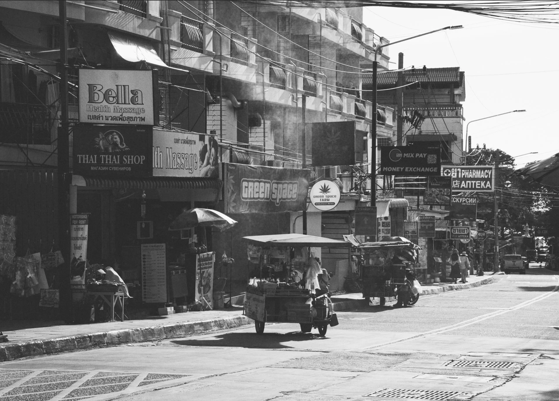 Street scene in an urban area with shops, signs, and a motorcycle. Small street vendors with carts line the sidewalk. Buildings have multiple signs, including a health massage, Thai shop, pharmacy, and money exchange. Utility poles and power lines ar