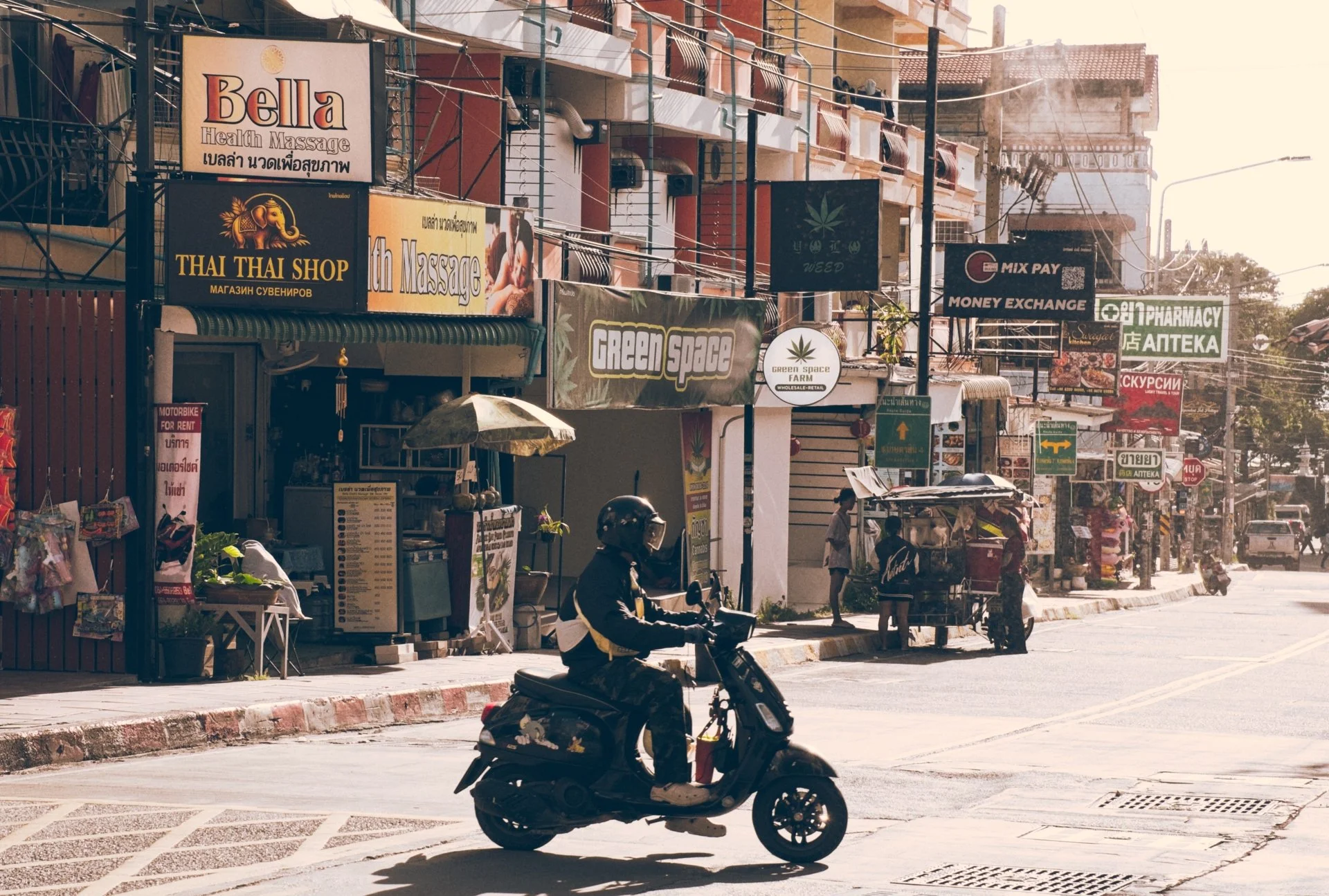 Street scene with signs for massage, Thai souvenir shop, weed, pharmacy, money exchange, and other businesses, with people on motorbikes and pedestrians on the sidewalk.