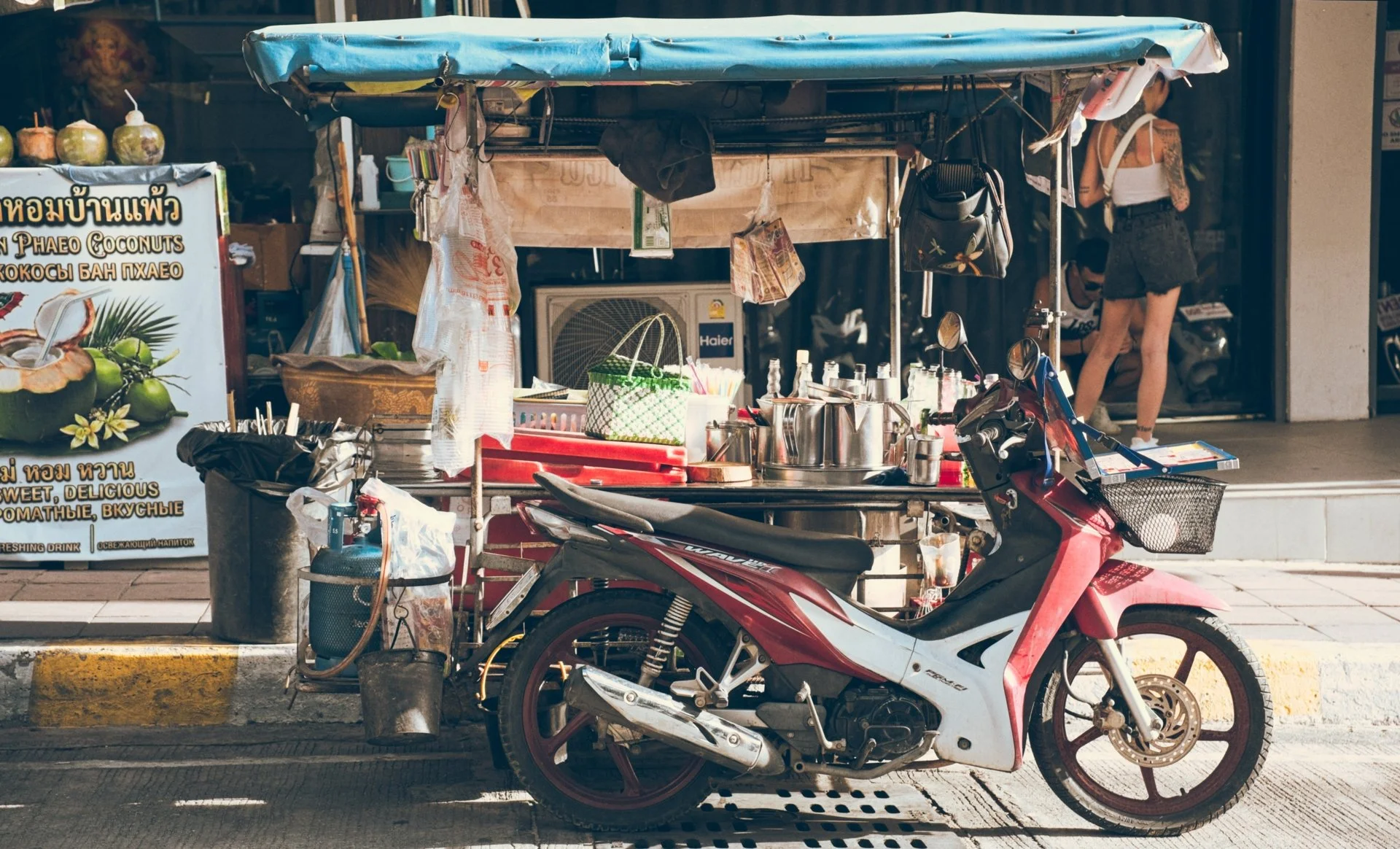 Street food cart with cooking utensils and a motorbike parked in front, advertisement sign on the side, and two women in the background near a storefront.