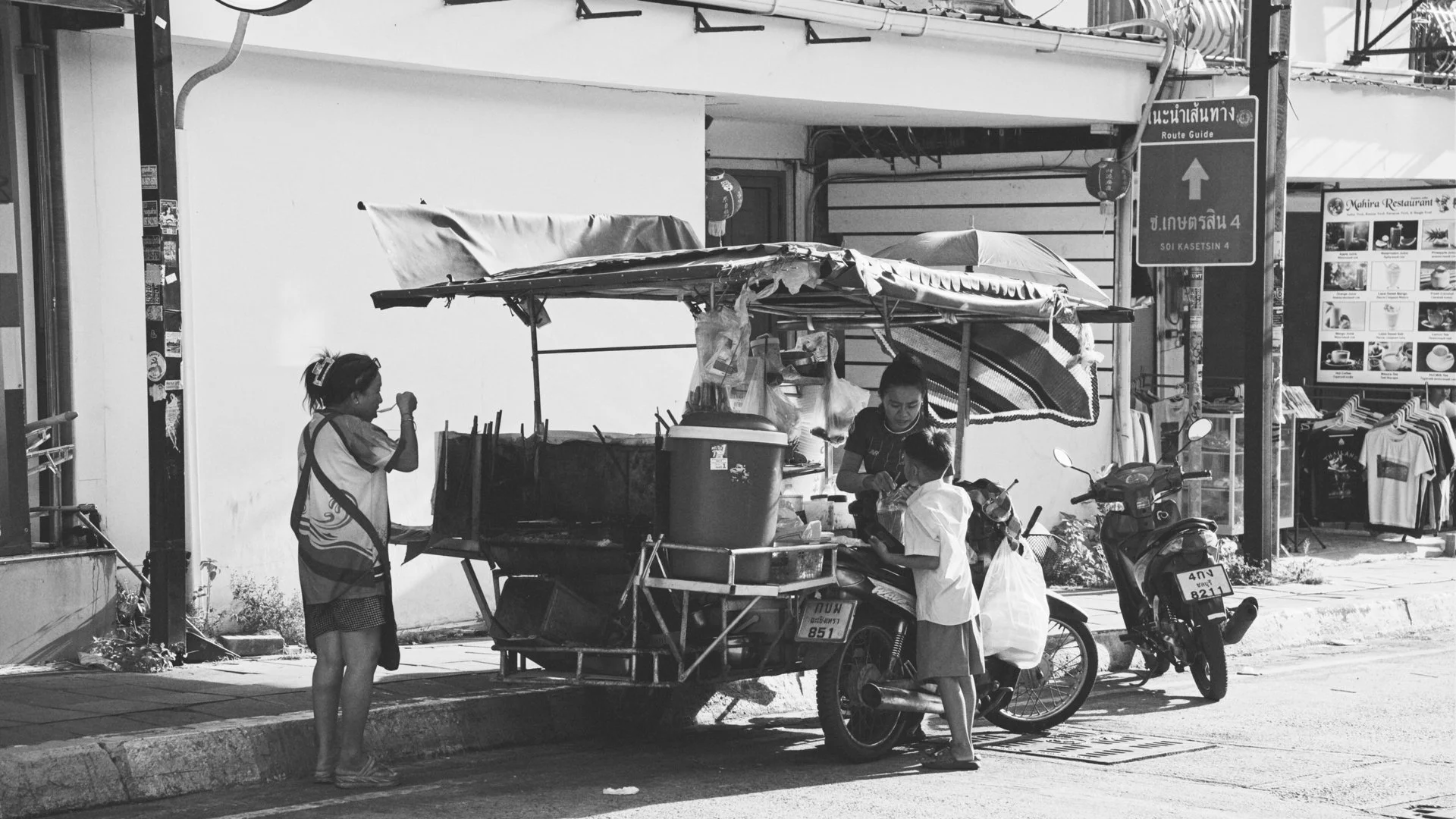 A street vendor setup with a small cart that has a canopy, where a woman is serving a boy, while another woman stands nearby. Two motorcycles are parked in front of the cart. There are street signs and a storefront with clothing on display in the bac