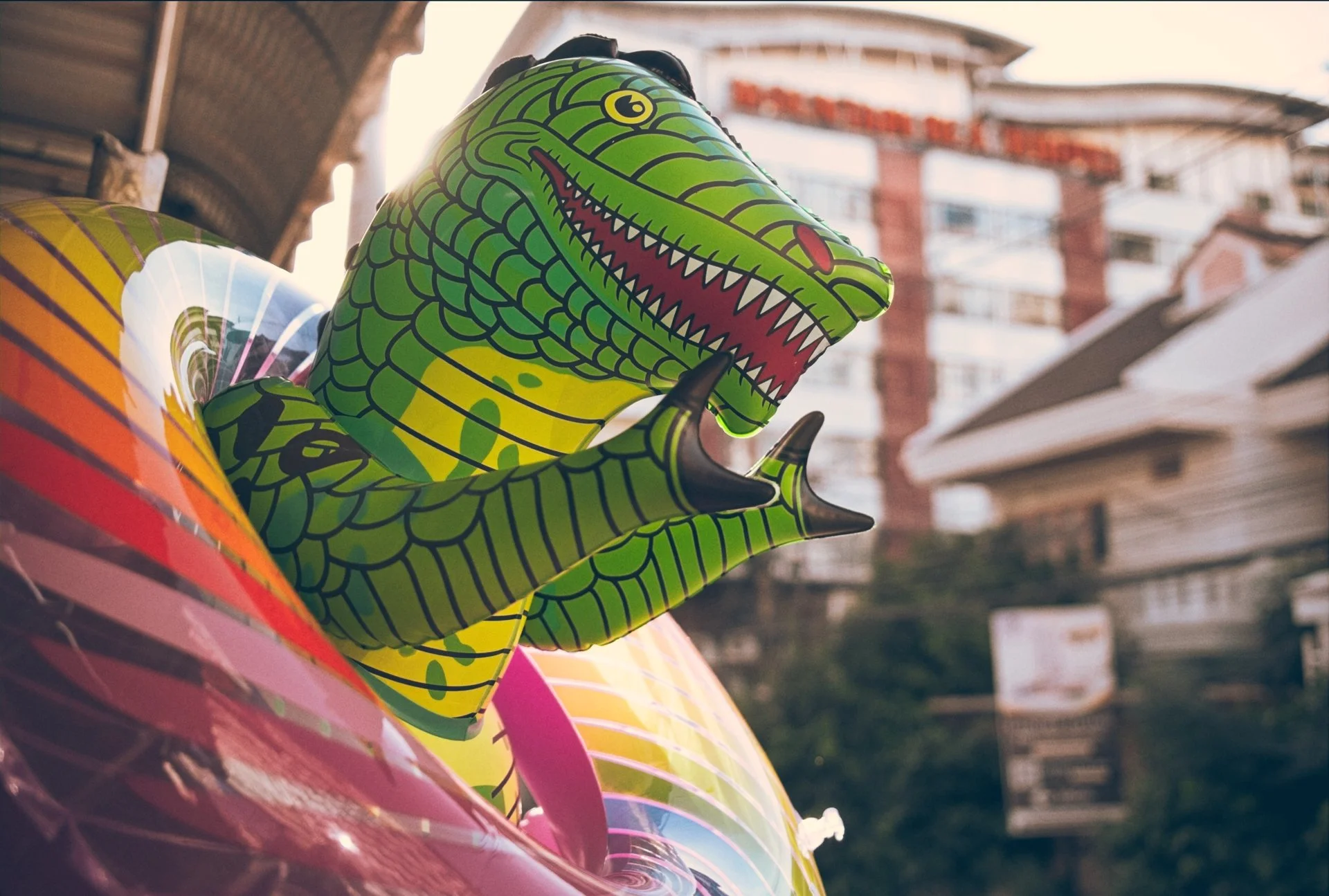 Close-up of a colorful inflatable crocodile toy, with a bright green body and detailed scales, featuring an open mouth with white teeth and a red tongue. The background shows a blurred cityscape with buildings and balconies.