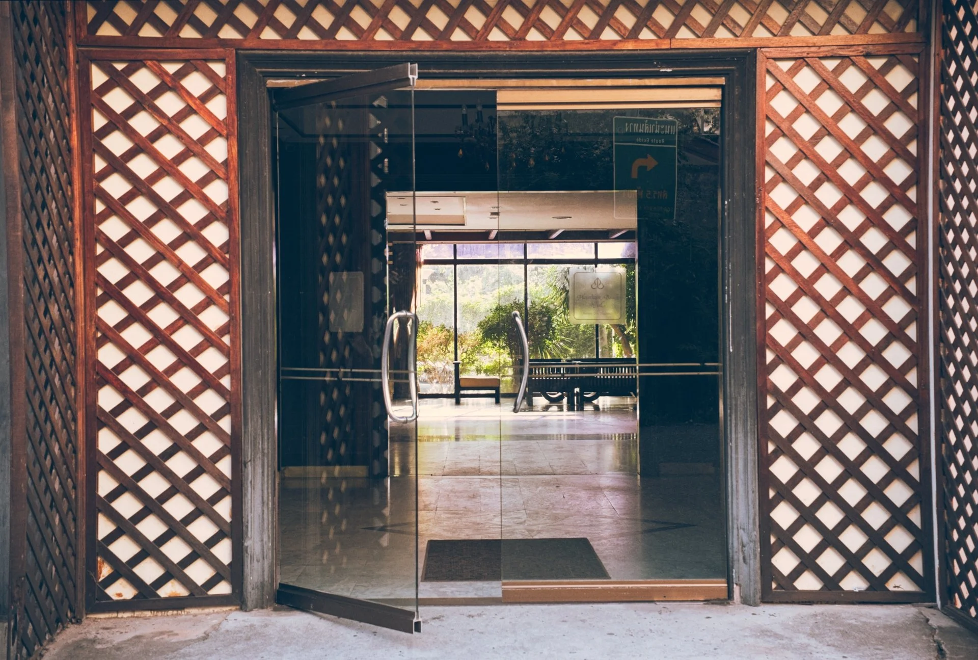 Glass door leading to an interior space with a large window and greenery outside, framed by wooden lattice panels.