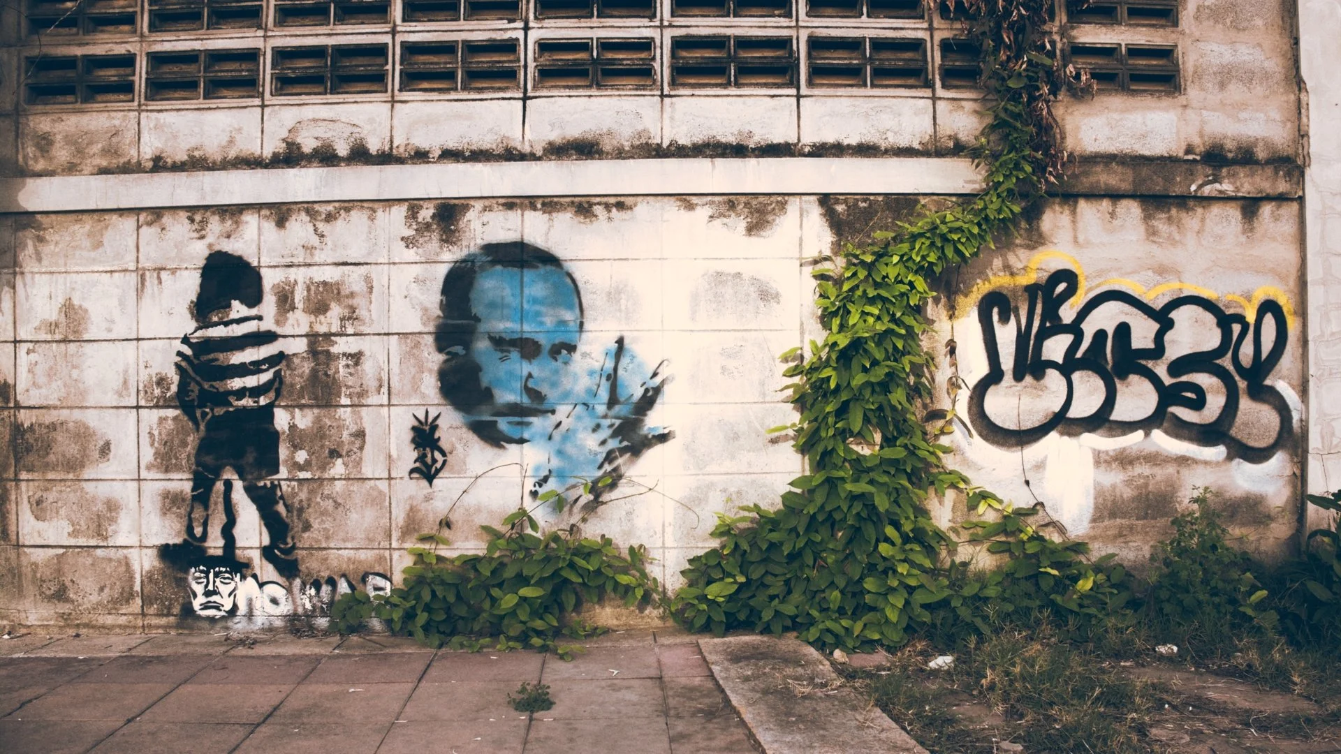 Street art graffiti on a weathered wall, featuring a blue portrait of a person with closed eyes and a relaxed expression, a smaller face at their feet, some black graffiti tags, and green climbing plants partially covering the wall.