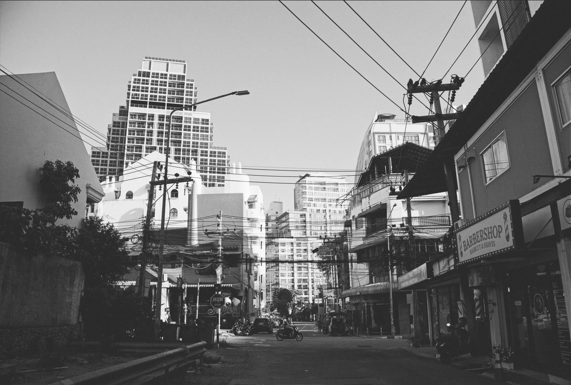 A black and white photo of an urban street scene with modern high-rise buildings in the background, utility poles and power lines overhead, and a few motorcycles and café signs at street level.