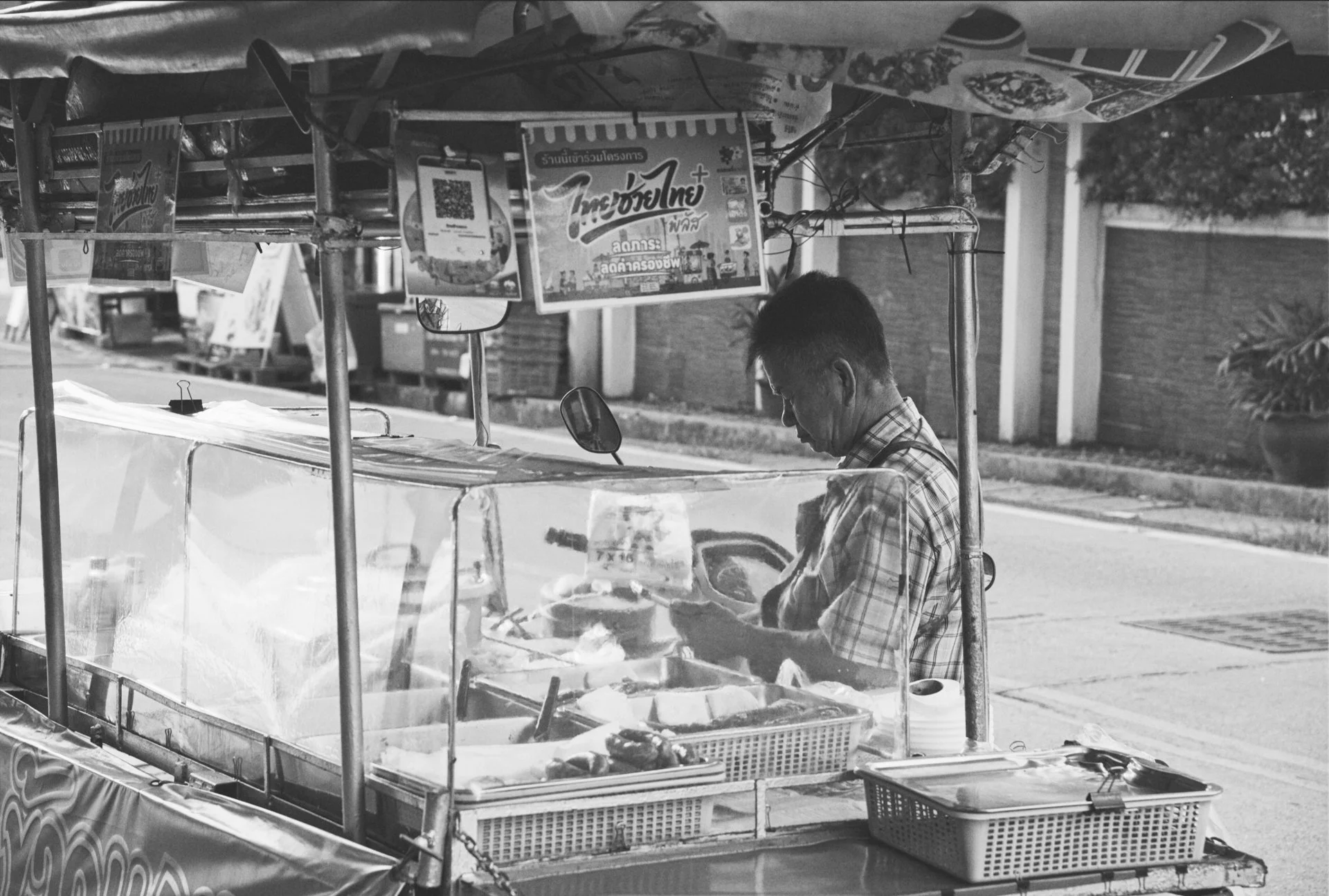 A street vendor selling food from a cart with clear plastic sides, situated on a sidewalk. The vendor is a man wearing glasses and a checkered shirt, focused on preparing food. Behind the cart, signage in Thai and various advertising posters are visi