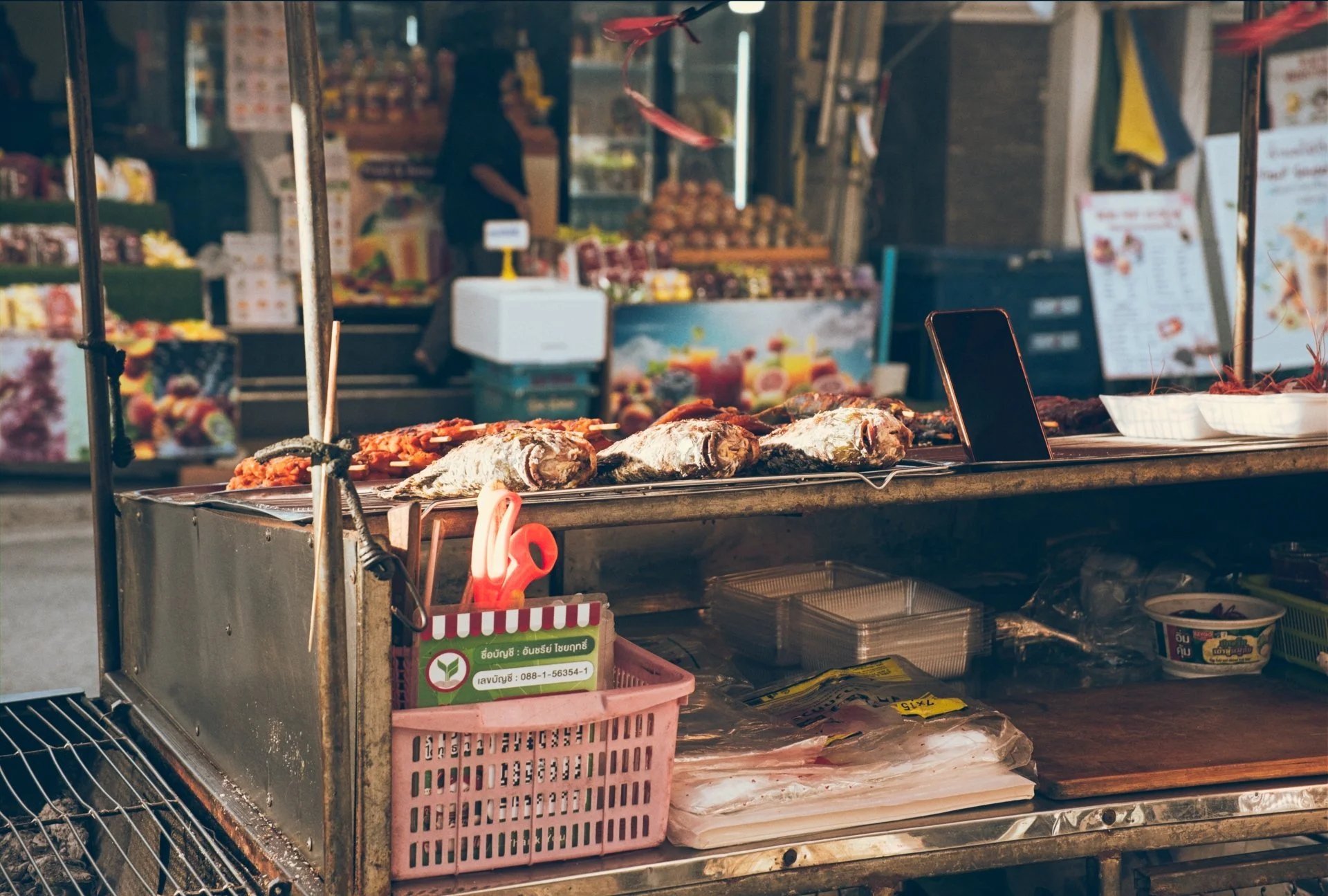 Street food cart with grilled fish, plastic containers, and a pink shopping basket in front, with a blurred market vendor and produce in the background.