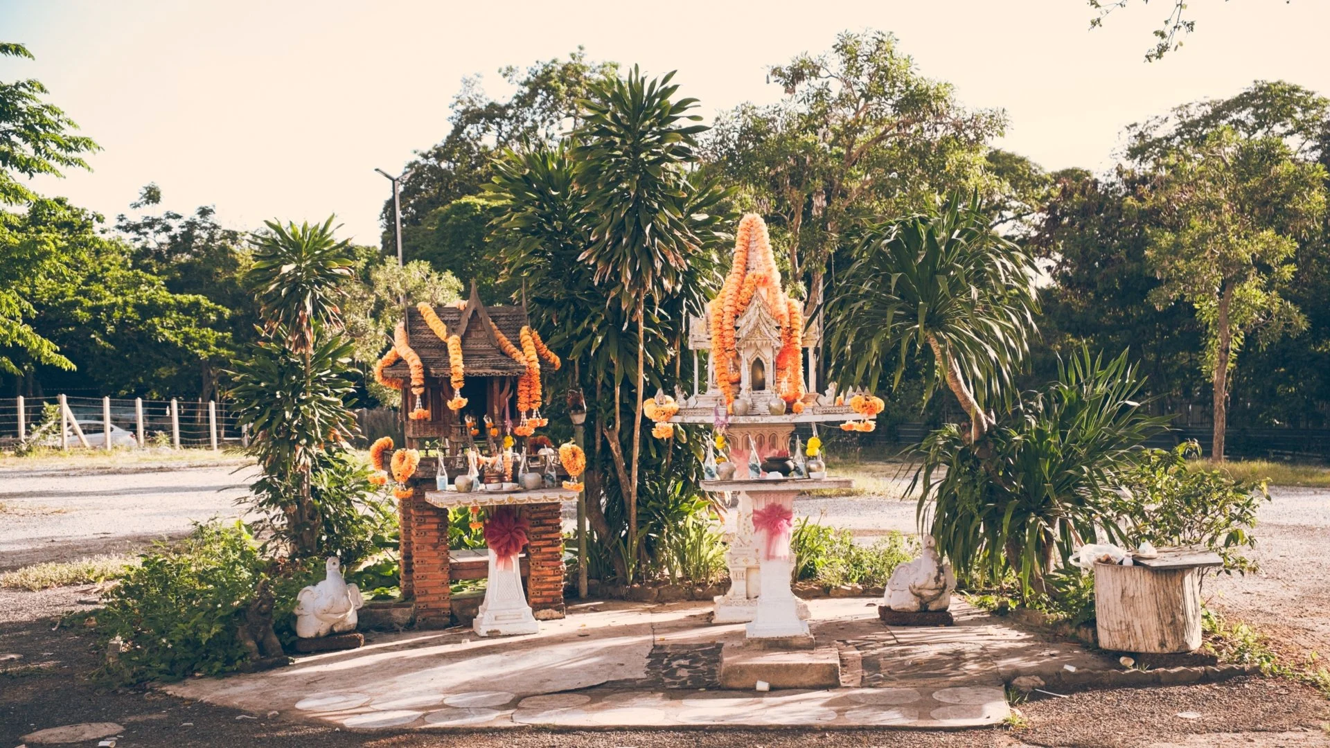 Small outdoor altar decorated with flower garlands and statues, set against lush green trees and a dirt road.
