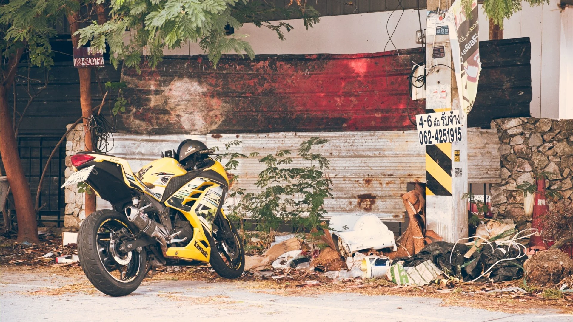 A yellow Kawasaki motorcycle parked on a street with scattered trash and debris, in front of a weathered, rusty metal shutter and a concrete wall with plants and trees around.