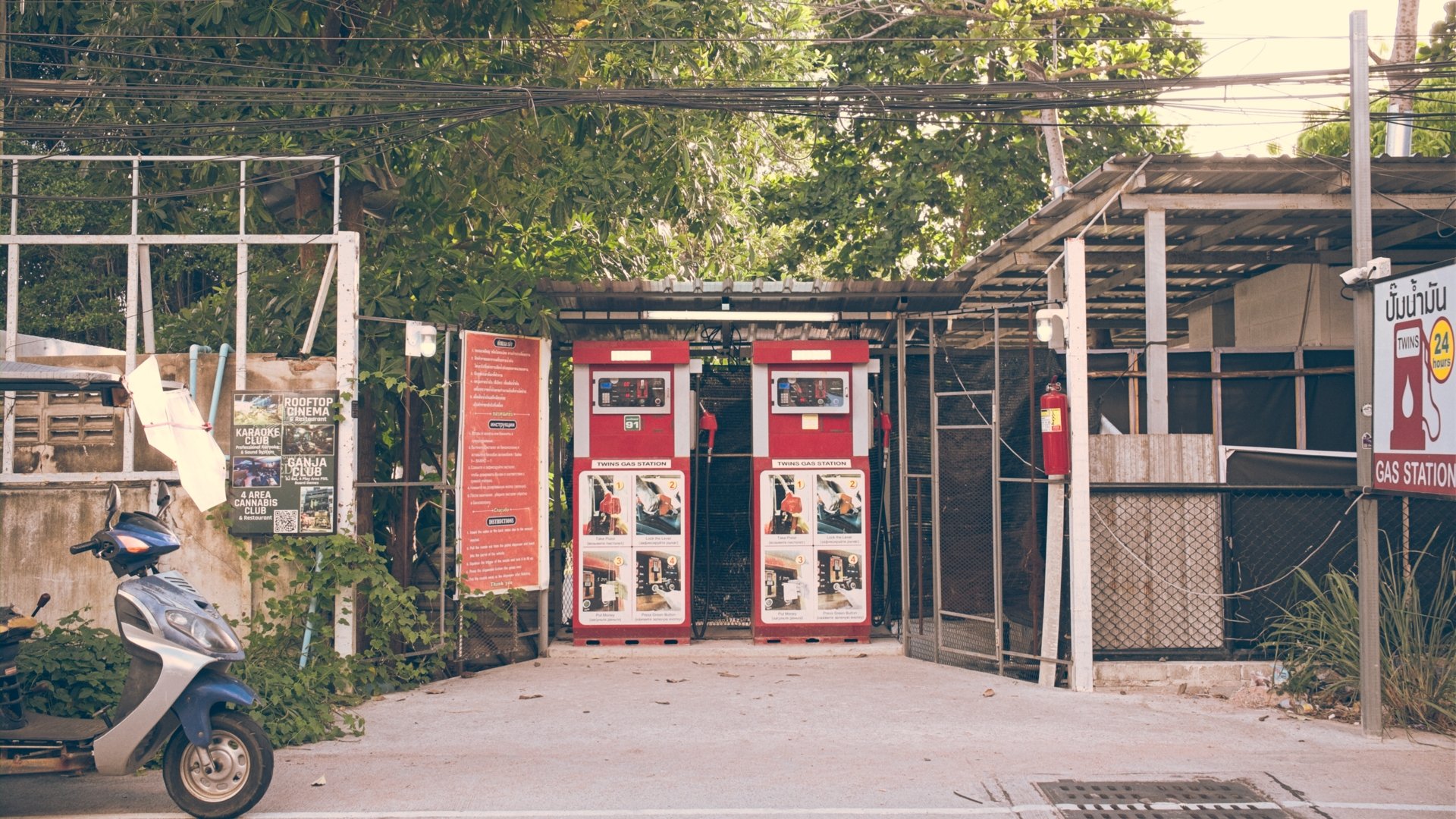 Two red gas pumps at a gas station with instructions displayed on a sign behind them. There are motorcycles parked nearby and trees in the background.