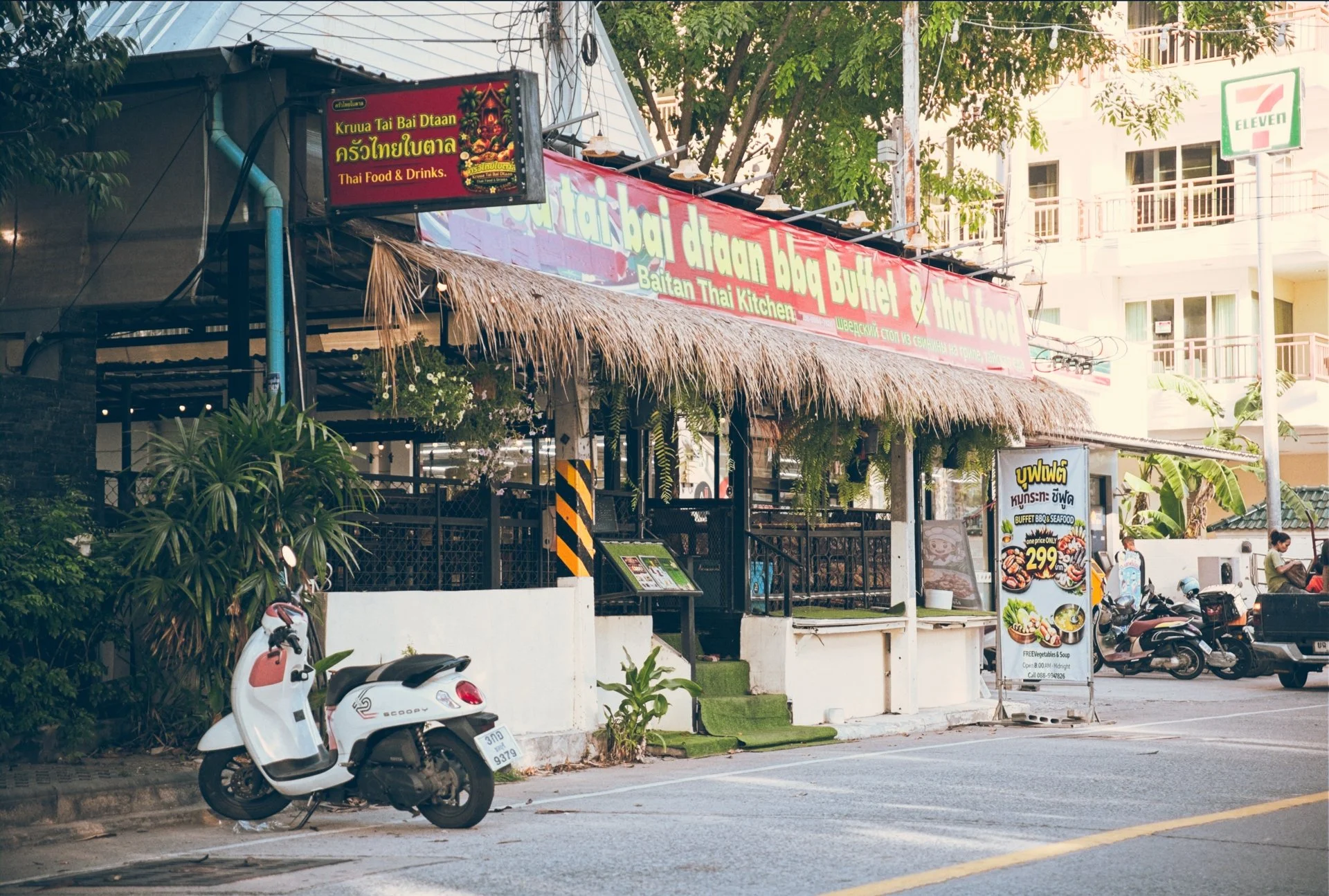 Street view of a Thai restaurant with a thatched roof and a sign with Thai and English text, parking scooters along the curb, and a 7-Eleven store sign in the background.