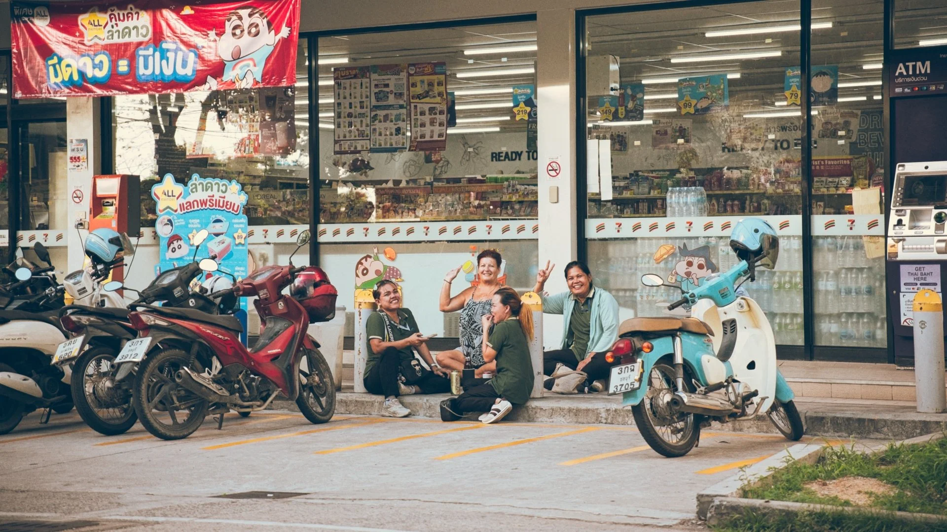 Four people sitting outside a convenience store, smiling and chatting, with motorbikes parked in front. The store has promotional posters and Thai language signs on the window.