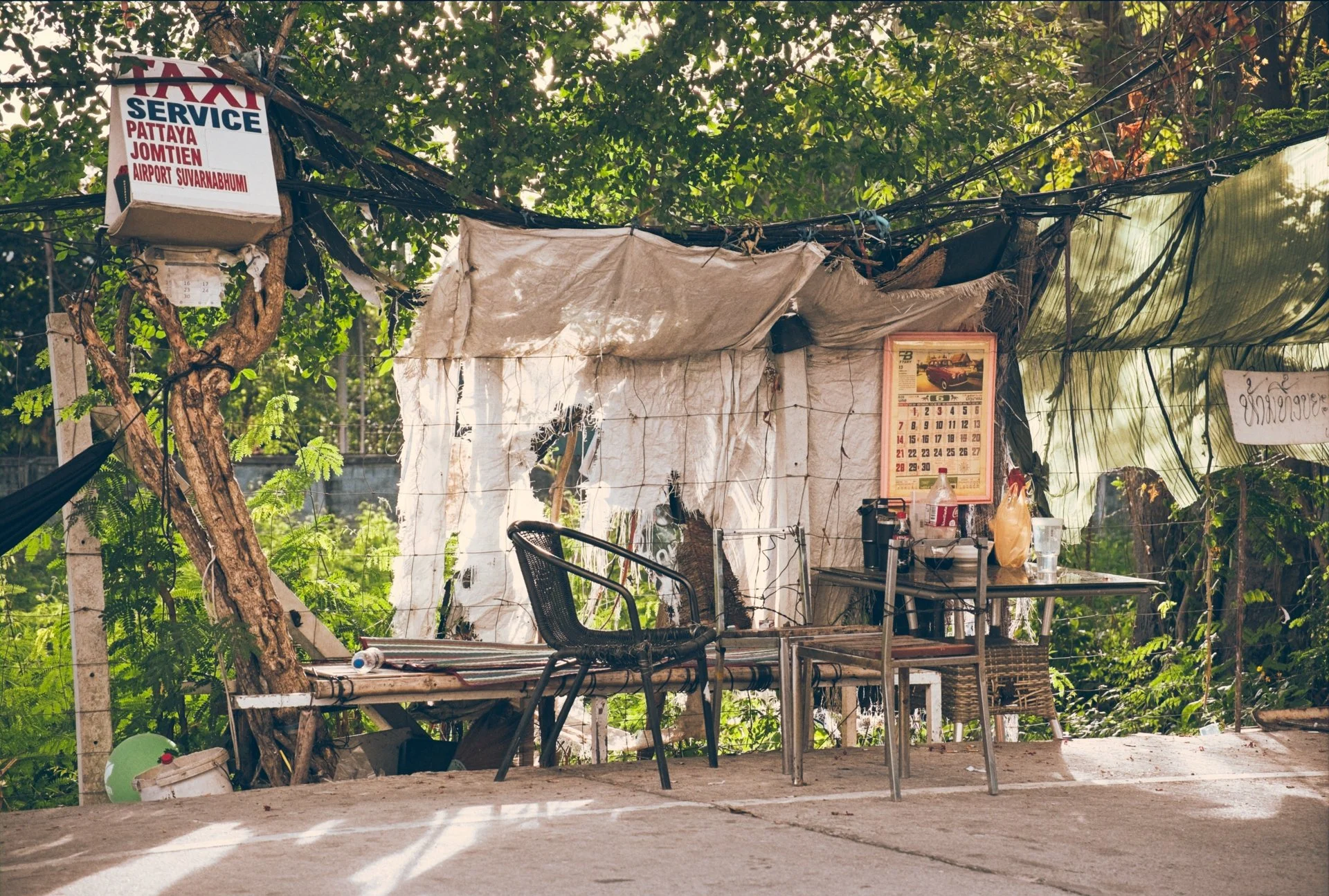A rustic outdoor seating area situated beneath a large leafy tree, featuring a simple metal table and chairs, with a worn fabric canopy and hanging electrical wires. There are various items on the table, including a plastic bottle, food, and containe