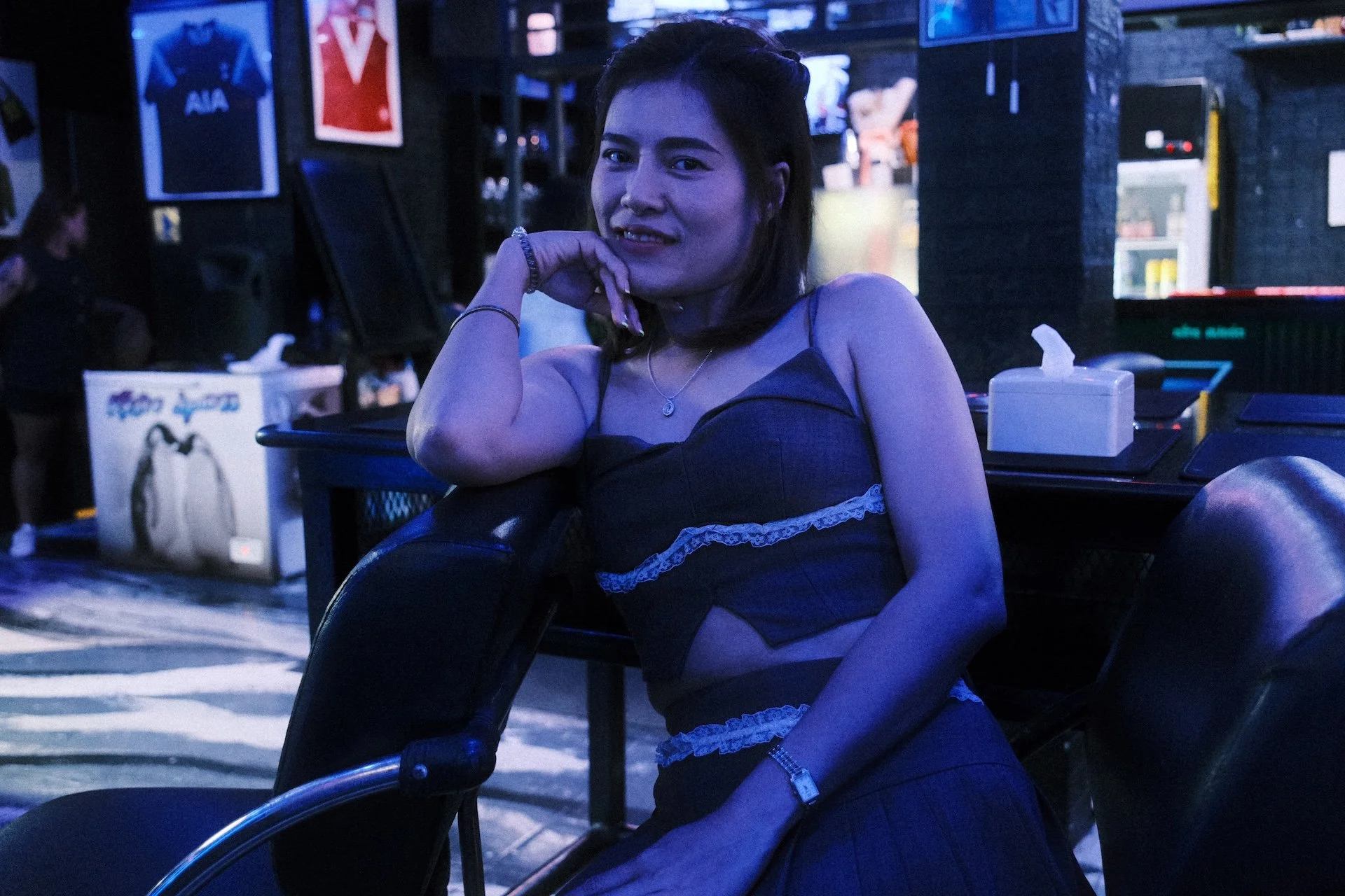A woman with shoulder-length dark hair and light skin sitting in a dimly lit indoor space, possibly a bar or nightclub, with her left hand resting on her chin. She is wearing a dark spaghetti strap top with lace details, a necklace, and a bracelet. I