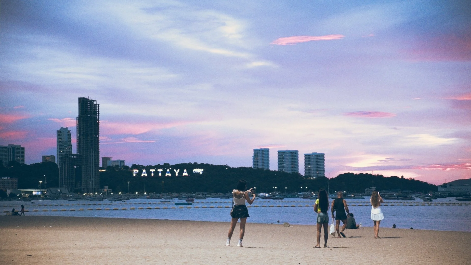 People on a sandy beach during sunset with high-rise buildings in Pattaya, Thailand, and the Pattaya sign in the background under a colorful sky.