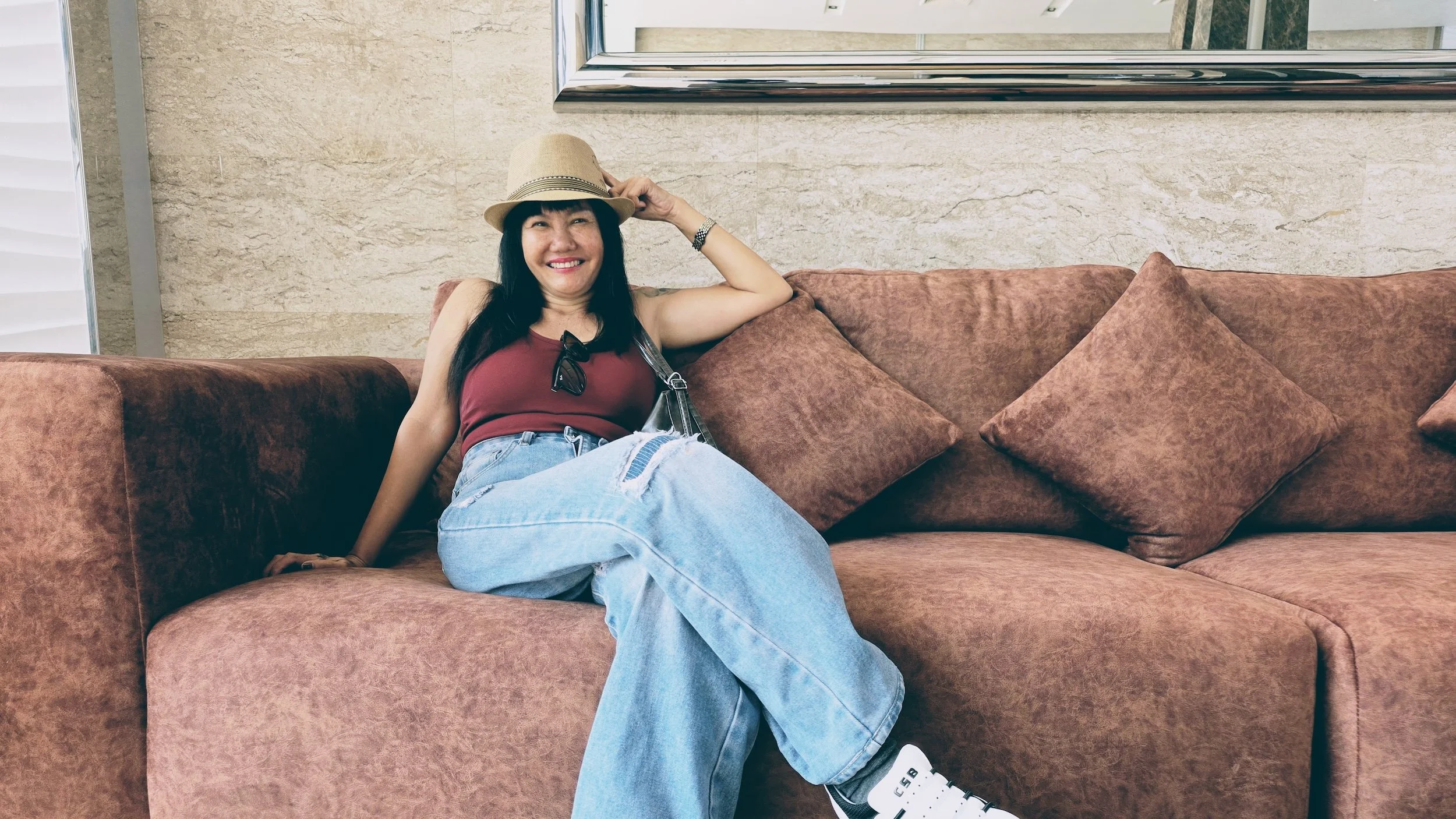 Smiling woman with long dark hair, wearing a straw hat, a red sleeveless top, and light blue ripped jeans, sitting on a brown couch with cushions in a modern interior.