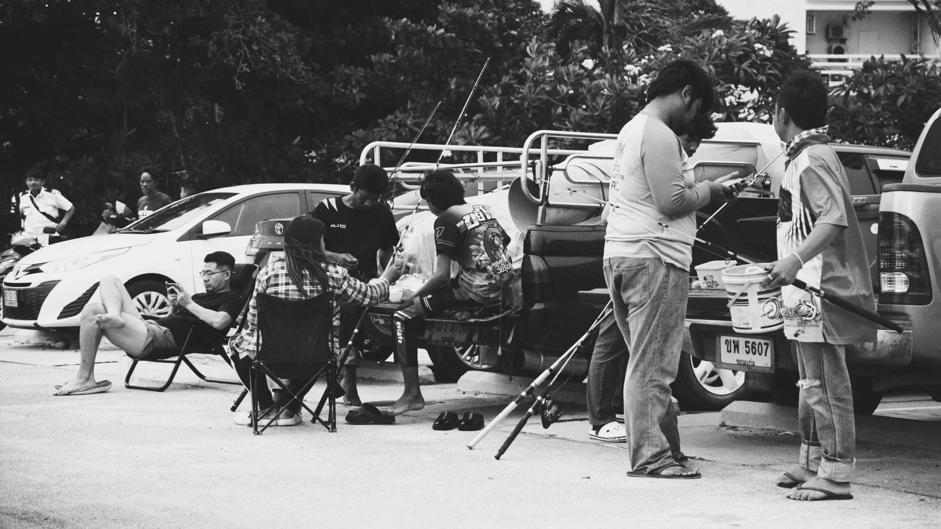 People fishing from a car parked in an outdoor parking lot, with some sitting on portable chairs and others standing, engaged in conversations or using their phones.