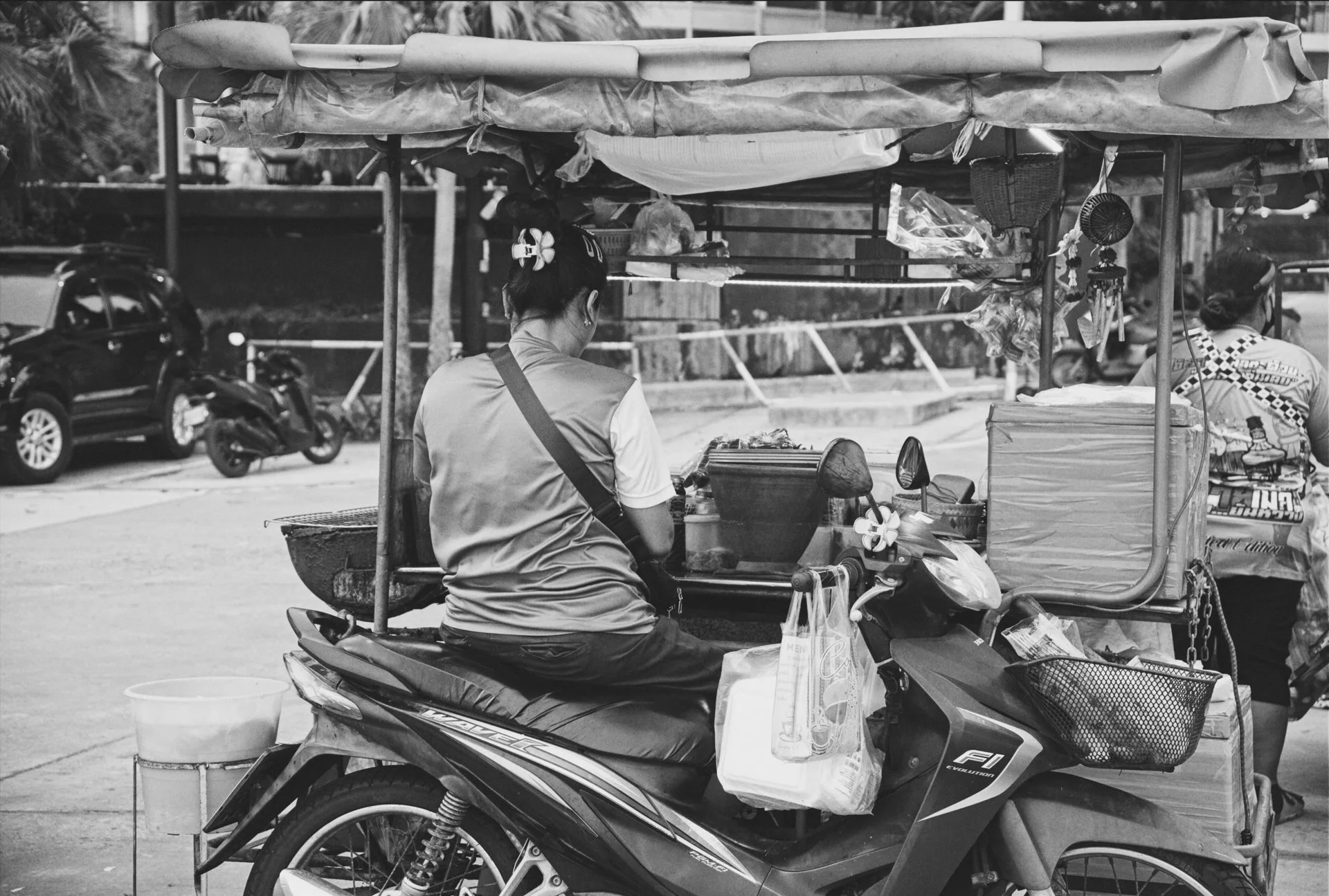 A woman operating a food or snack delivery scooter with a cart attached, parked on a city street. The cart is filled with supplies and snacks, and there are other parked vehicles and pedestrians in the background.