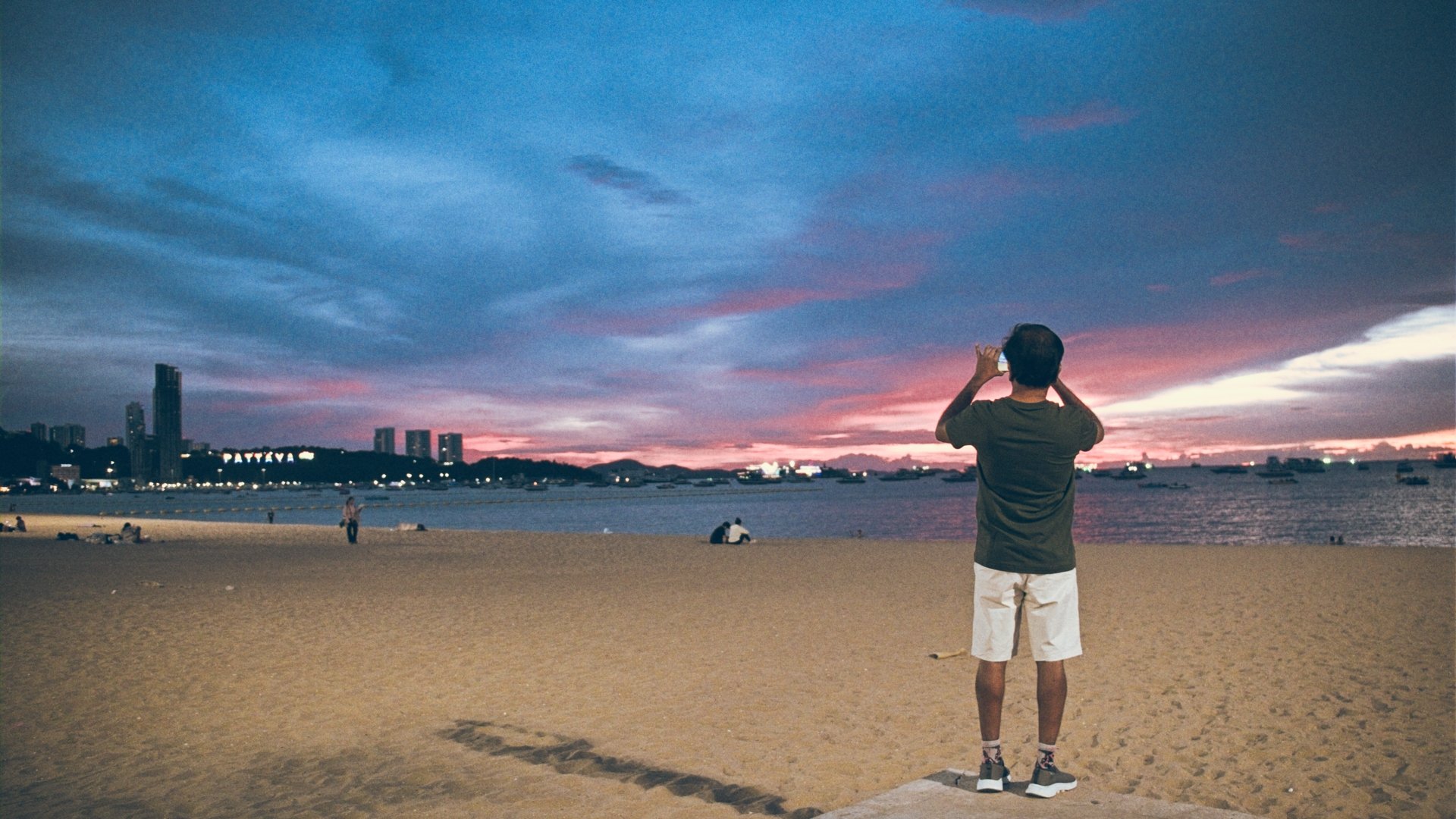 A person standing on a sandy beach captures the colorful sunset sky with a smartphone, with a city skyline and boats on the water in the background.