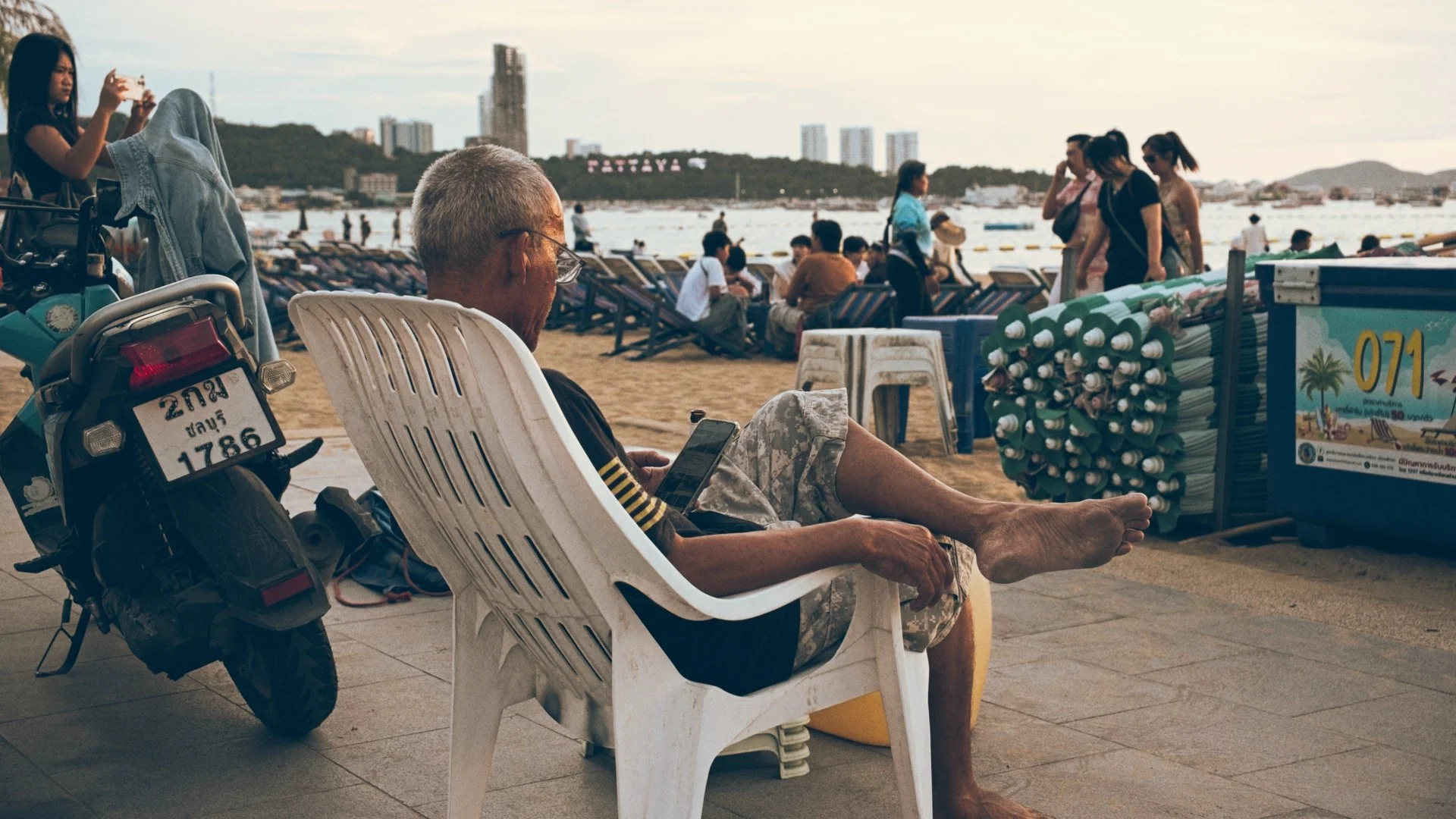 An elderly man with gray hair and glasses sitting on a white plastic chair, relaxing on a beachside promenade, looking at his phone. A motorcycle with a Thai license plate is parked beside him. In the background, there are people at the beach, some s