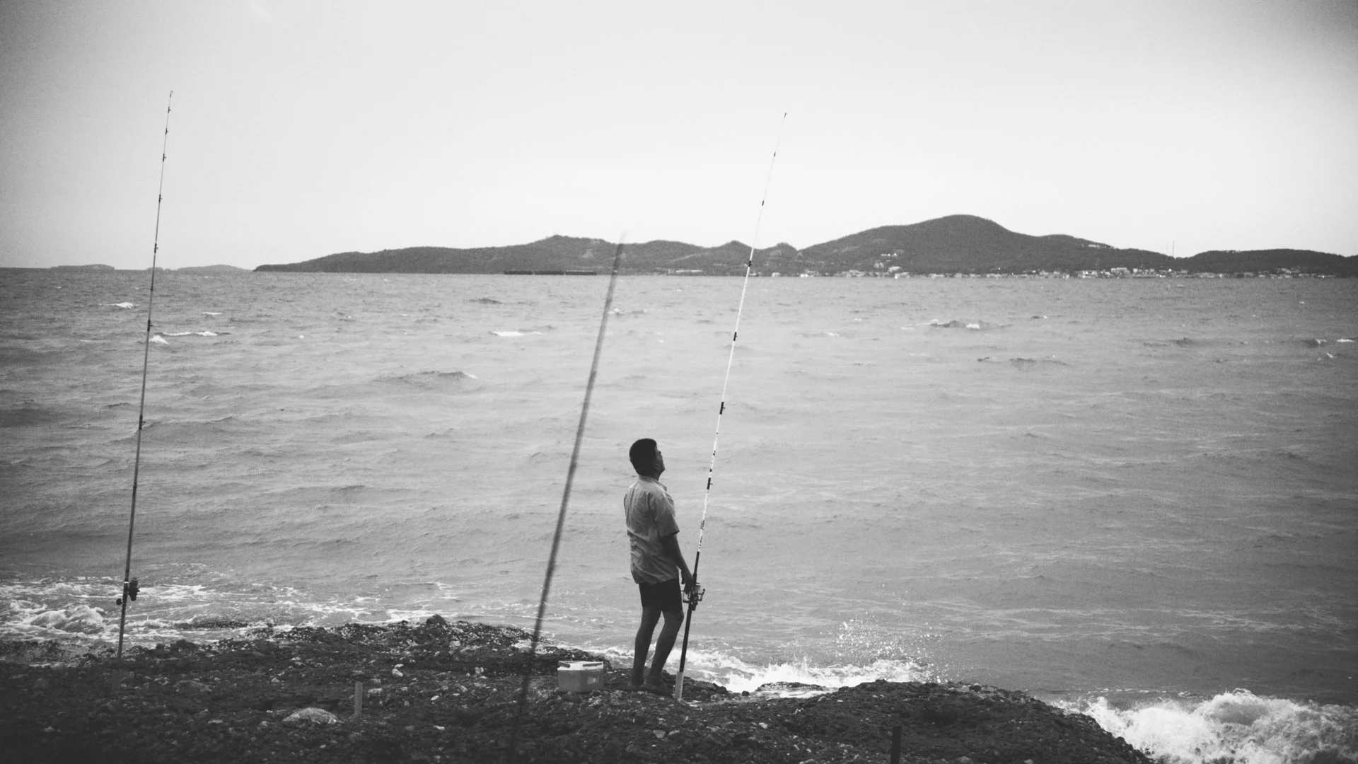 A man standing on a rocky beach, fishing with two fishing rods, with a lake or ocean and distant hills in the background, black and white photo.