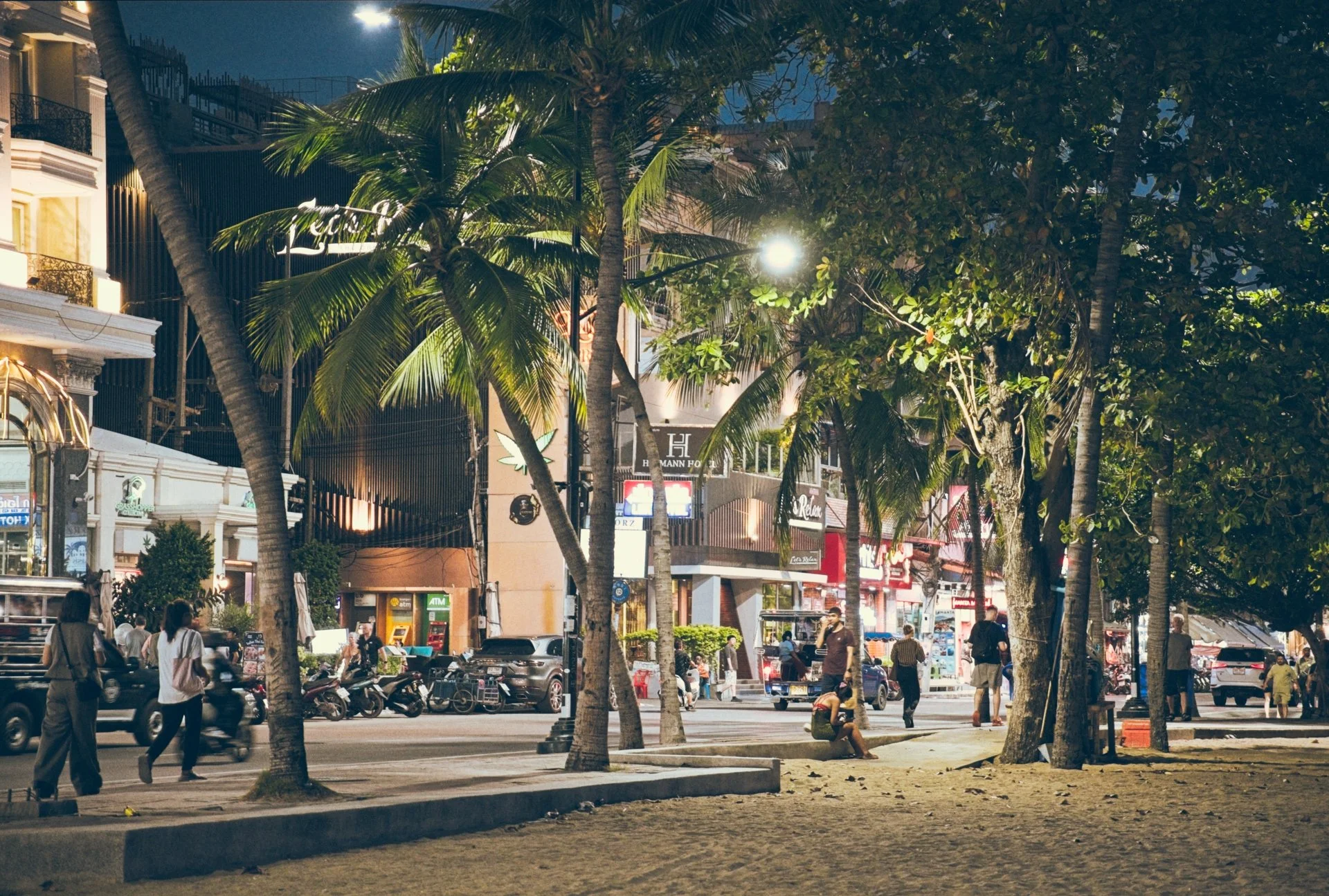 A vibrant city street scene at night with palm trees and lush green foliage in the foreground. People are walking and sitting, and various businesses with neon signs and storefronts line the street. The scene is illuminated by streetlights and buildi