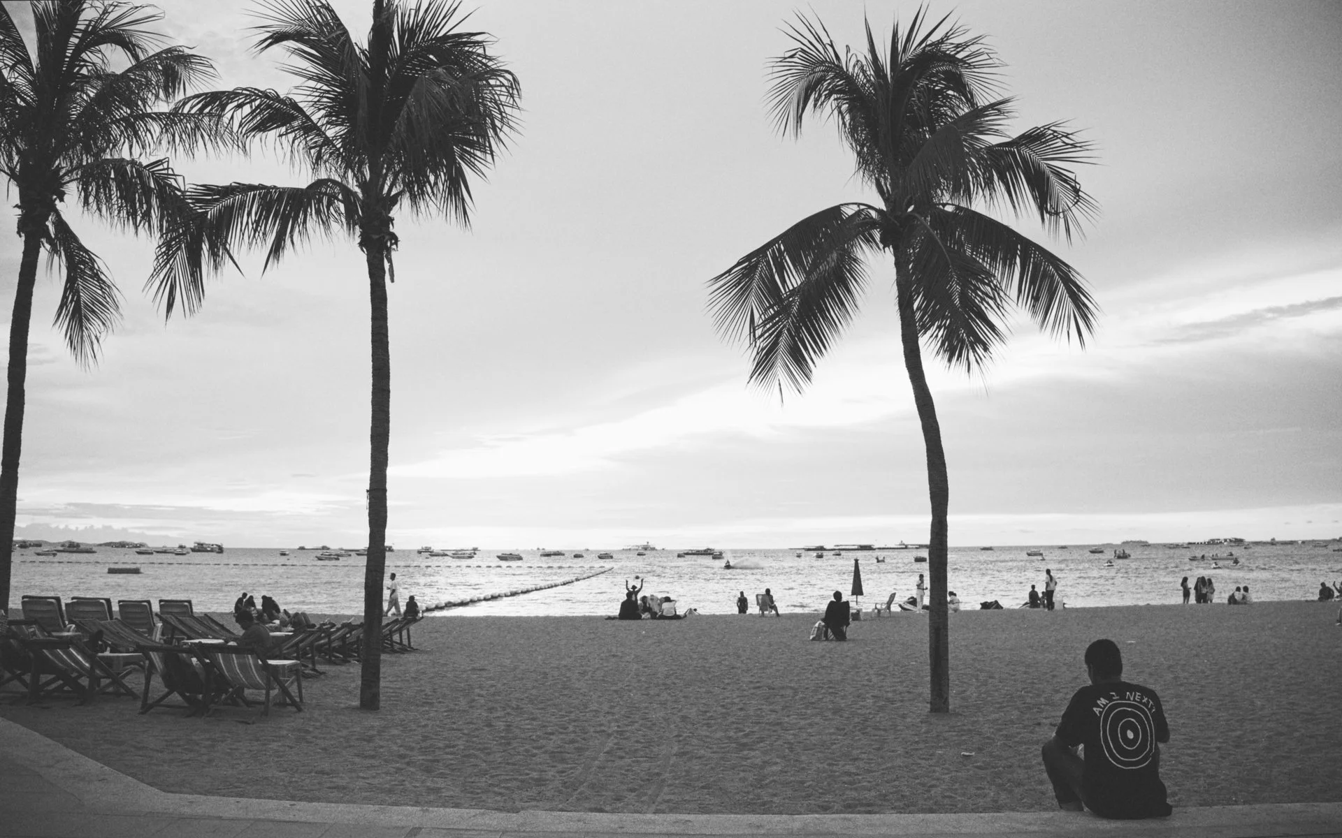 Black and white photo of a beach scene with two tall palm trees in the foreground. People are sitting and walking on the sandy beach, some by lounge chairs, and boats are anchored in the water in the background. Clouds are visible in the sky.