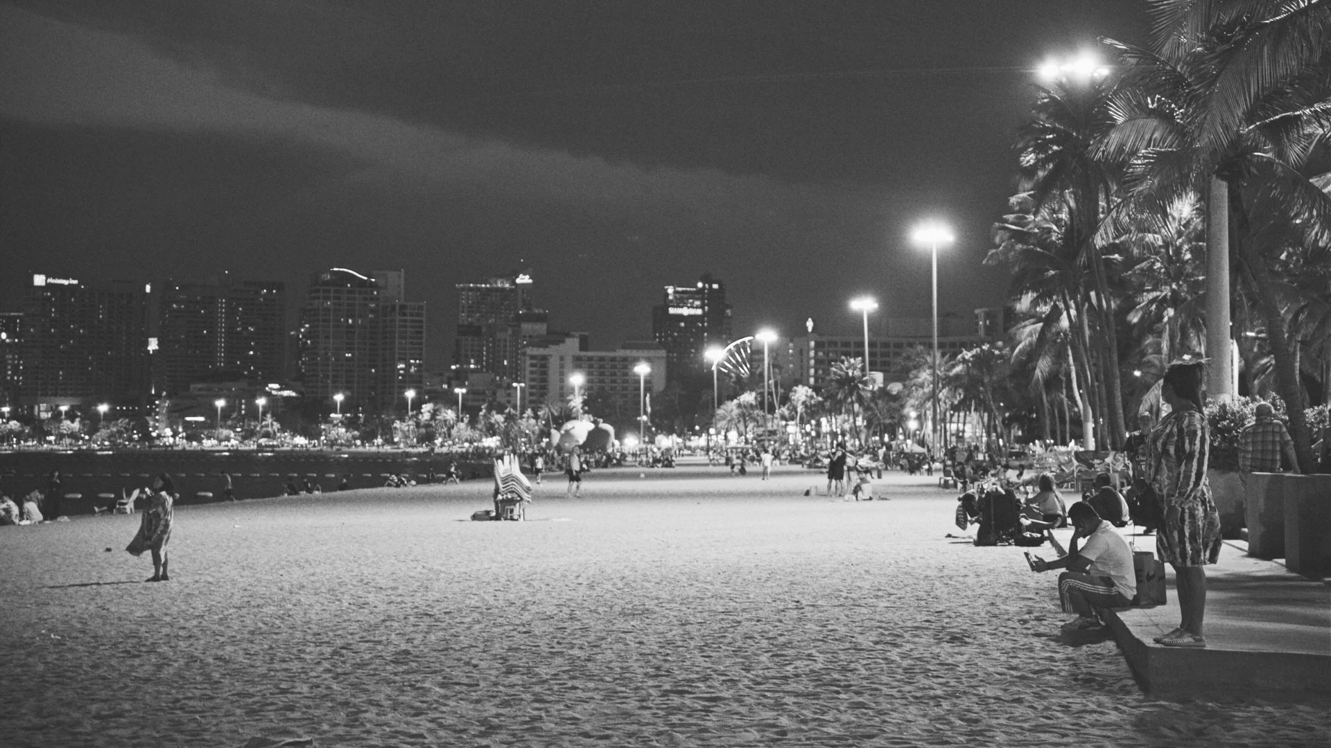 Nighttime scene at a beach with people sitting and walking, palm trees illuminated by streetlights, and a city skyline with tall buildings in the background.