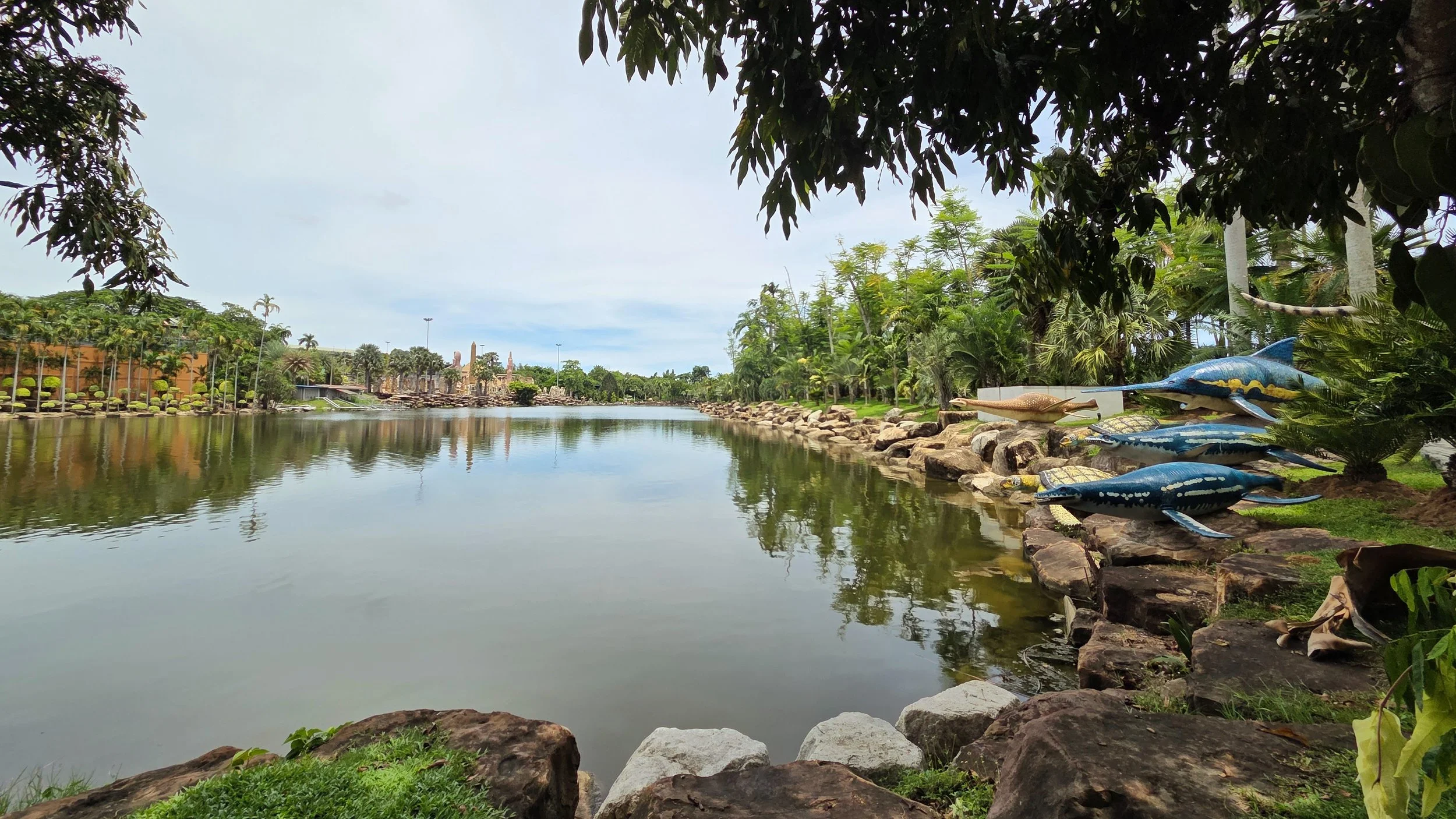 A serene body of water reflecting a cloudy sky, surrounded by lush green trees and decorative crocodile sculptures along the rocky shoreline.