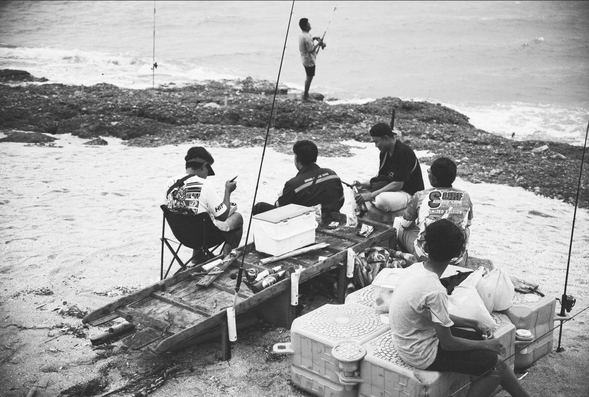 Group of people fishing and relaxing by the rocky shore. Some are sitting on coolers, while others are standing and fishing with rods. The scene is in black and white.