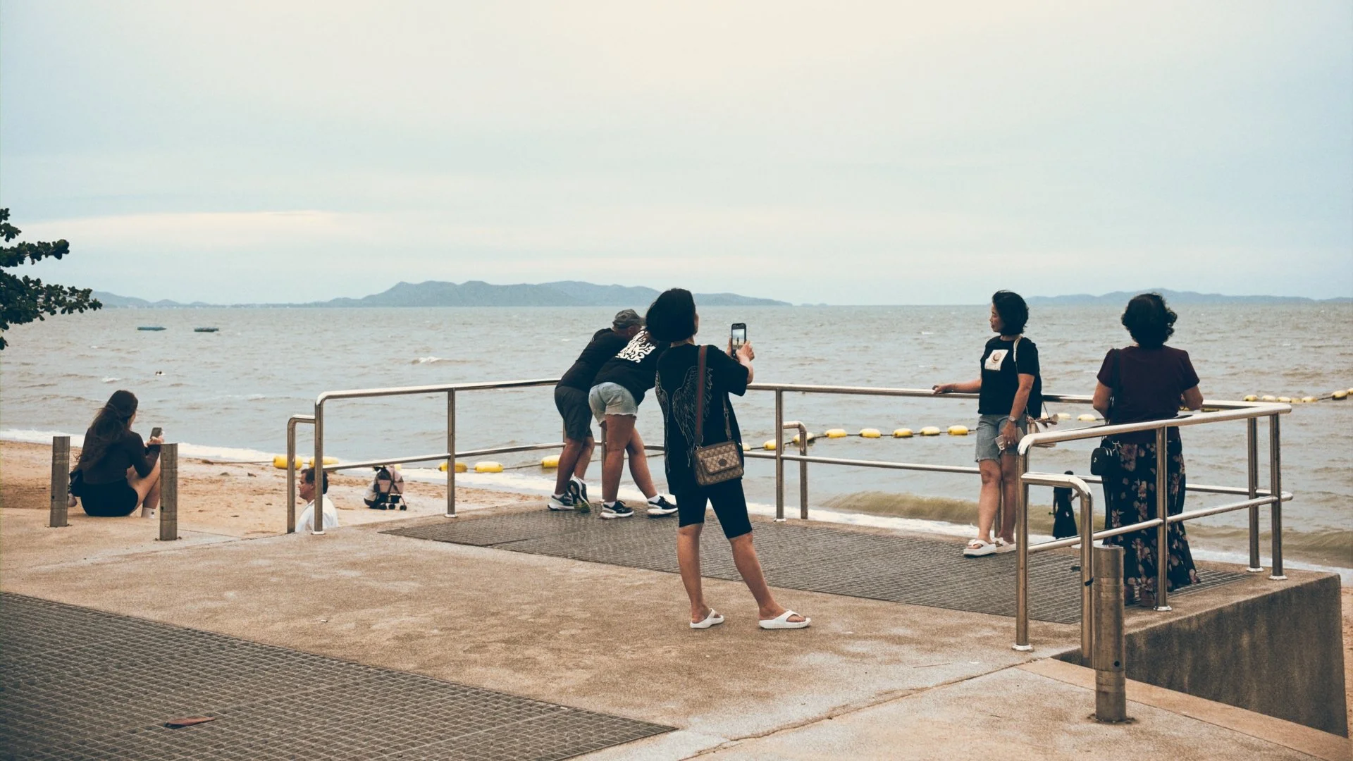 People taking photos and enjoying the view of the water from a seaside promenade with railings and a distant island or shoreline in the background.