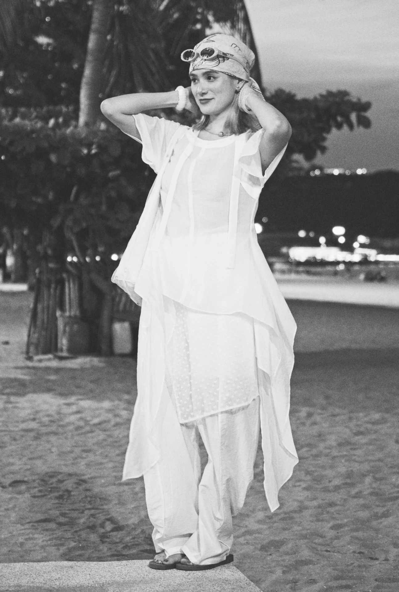 A woman standing outdoors on a beach at dusk, wearing a flowing white dress, a headscarf, and goggles, with her hands behind her head and smiling.