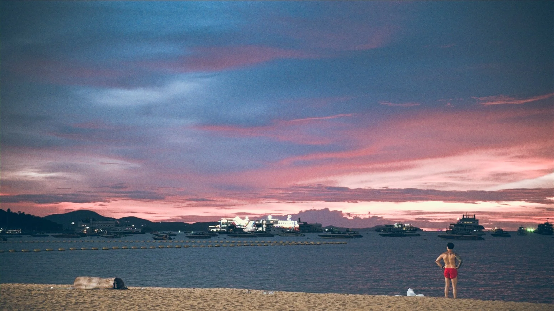 A person standing on a sandy beach at sunset, with boats on the water and a colorful sky with pink, purple, and blue clouds.