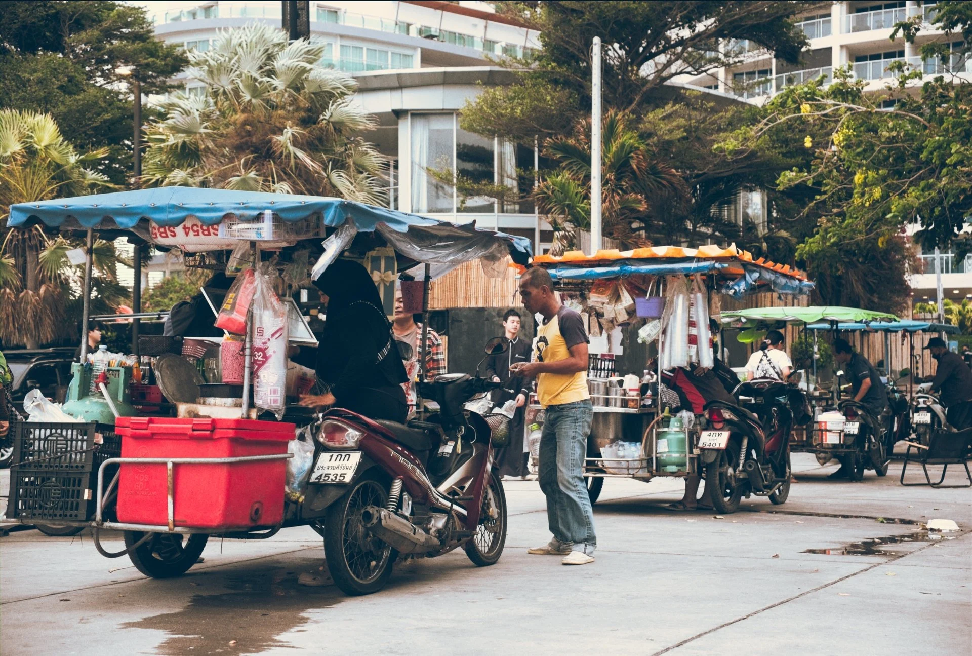 Street market scene with vendor stalls under blue and orange canopies, motorcycles parked nearby, and people shopping and selling, with modern buildings and trees in the background.