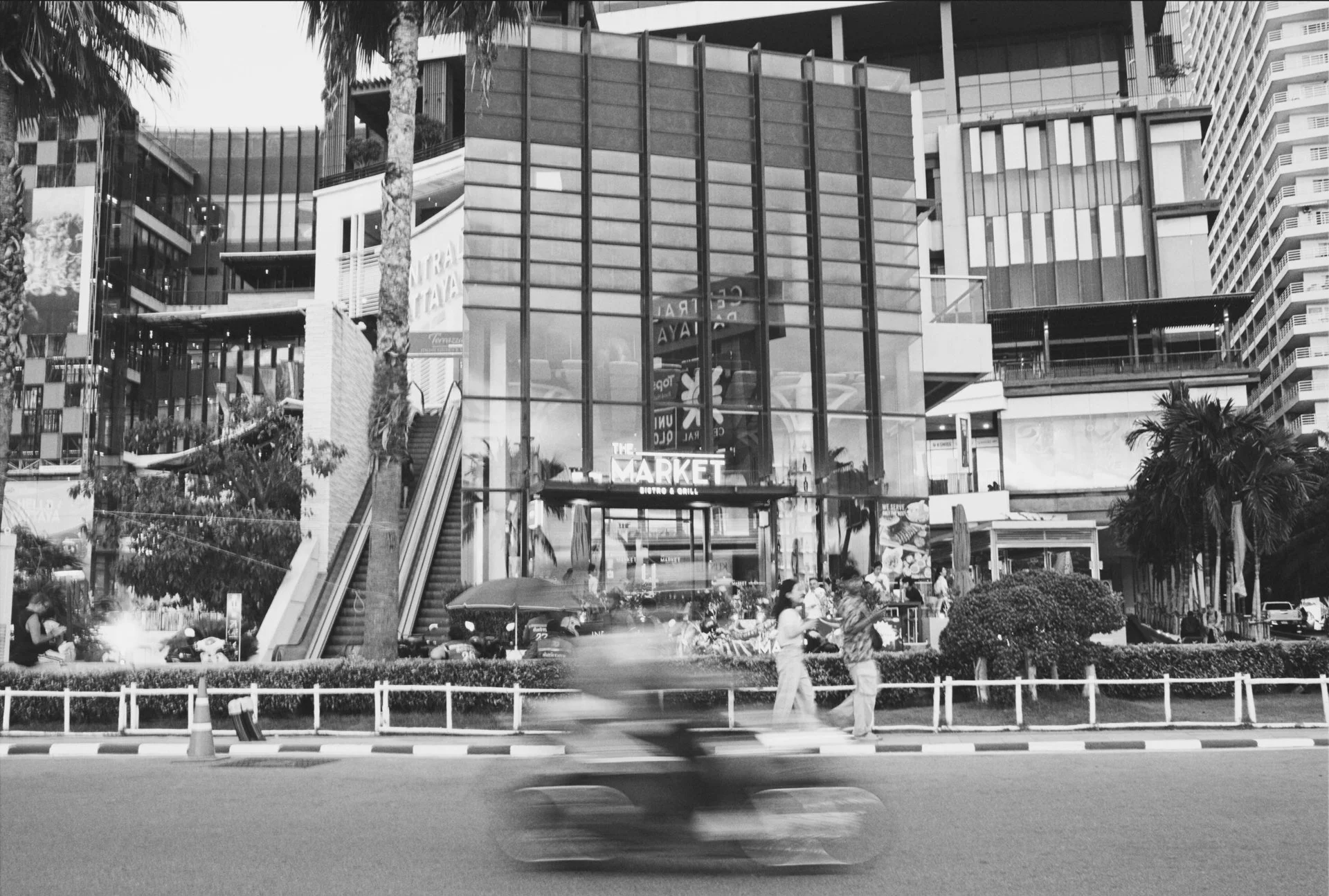 A bustling city scene featuring a modern glass-fronted building with a sign reading 'THE MARKET' at its entrance. There are escalators leading up to the building, tall trees, and people walking in the foreground as a blurred bicycle passes by, indica