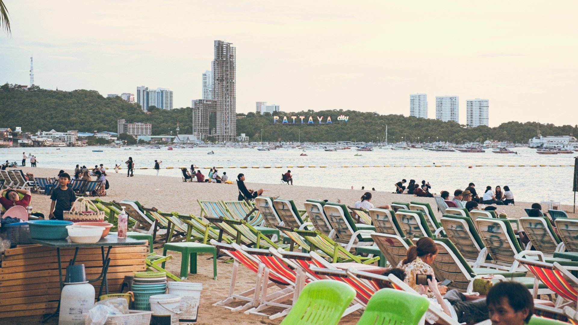 Beach scene with people sitting and relaxing on chairs, some standing or walking, with a view of the city skyline and boats on the water in Pattaya.