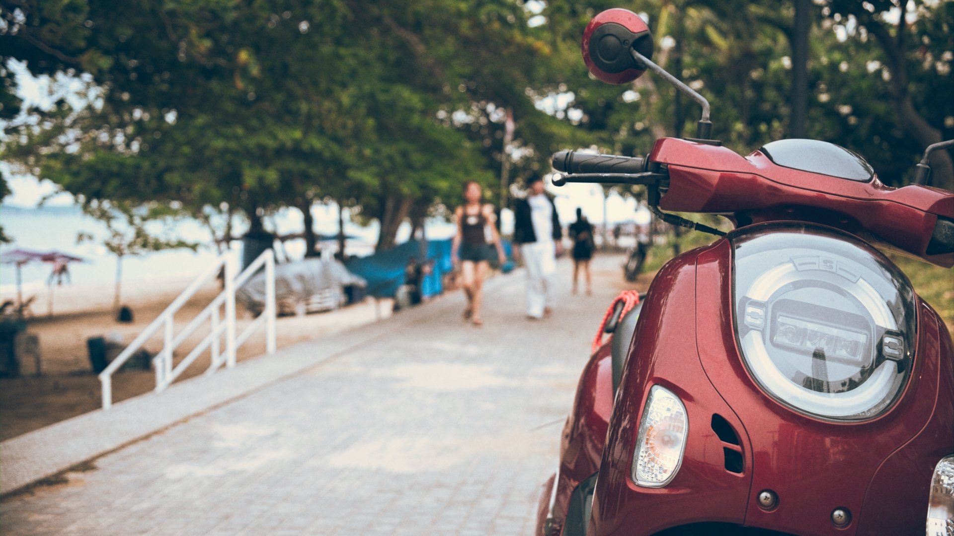 Close-up of a red electric scooter on a sidewalk with blurred people walking in a park with trees and a beach in the background.