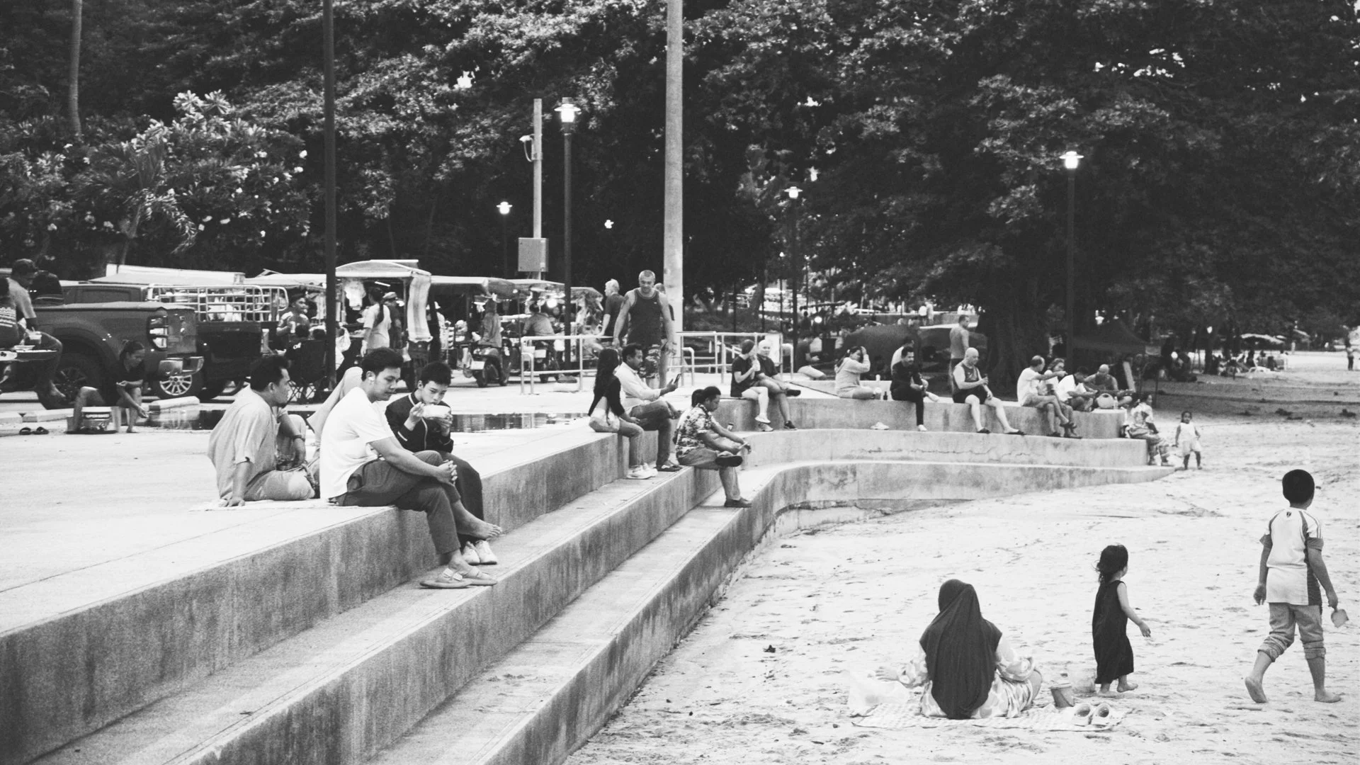 People sitting and walking along concrete steps at a park or beach area with trees and streetlights in the background.