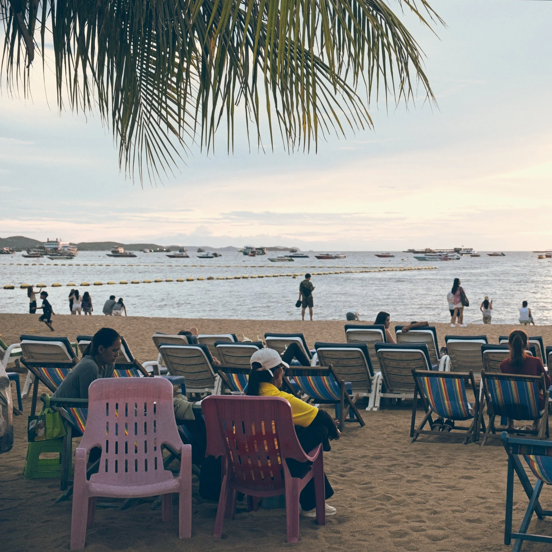 Beach with colorful plastic chairs, people walking and sitting by the water, boats anchored in the ocean, palm tree branch overhead, cloudy sky.