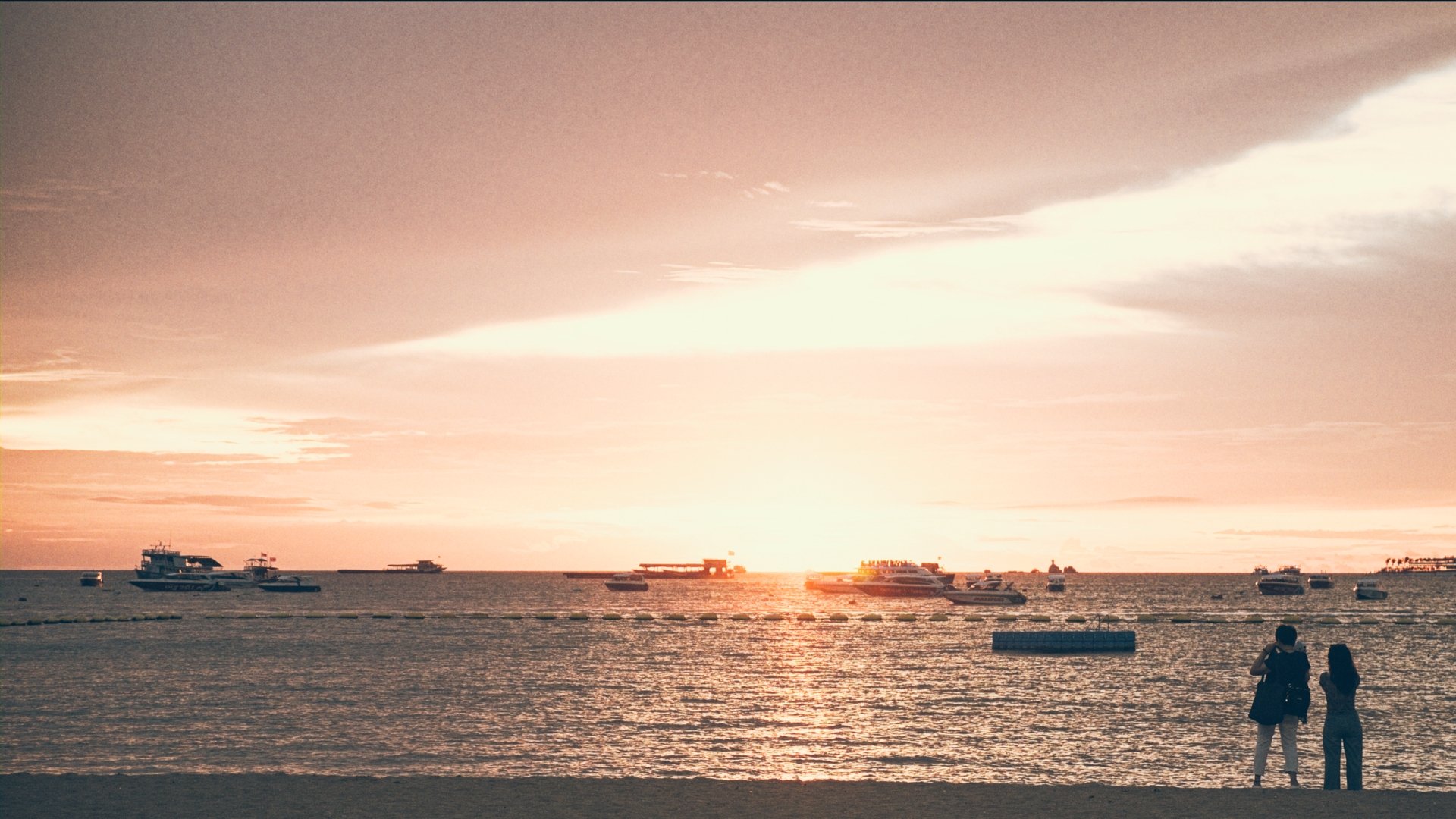 A sunset over the ocean with boats anchored near the shore and two people standing on the beach watching the scene.