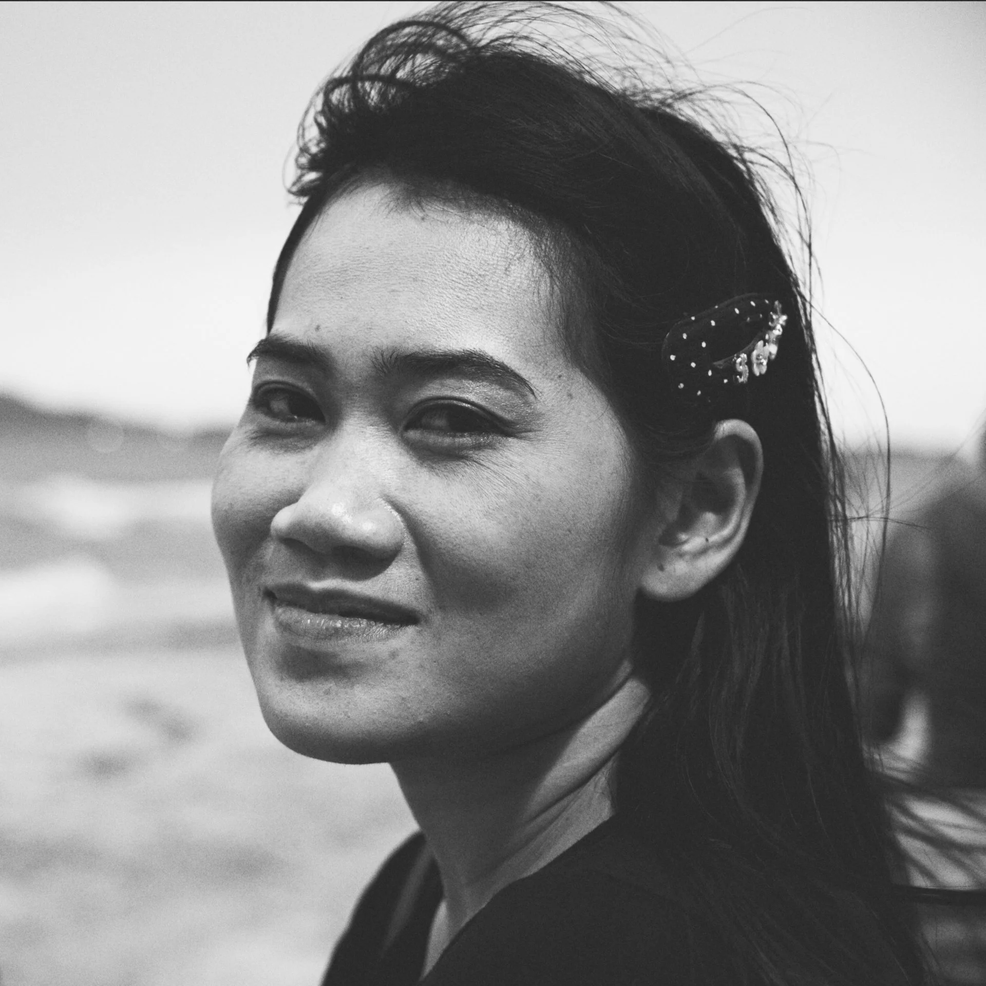 A black-and-white close-up photograph of a smiling woman with dark hair, wearing a hair clip adorned with small pearls and rhinestones.
