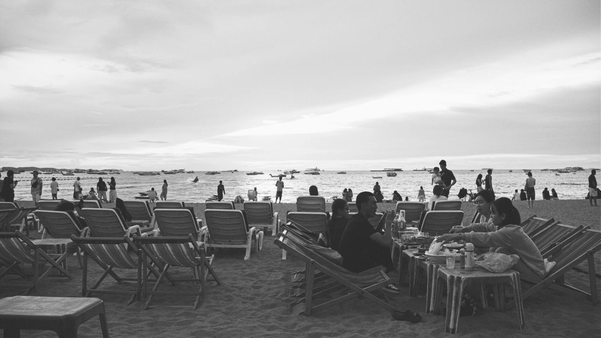 People sitting and standing on a beach with lounge chairs facing the ocean, boats on the water, and a cloudy sky.