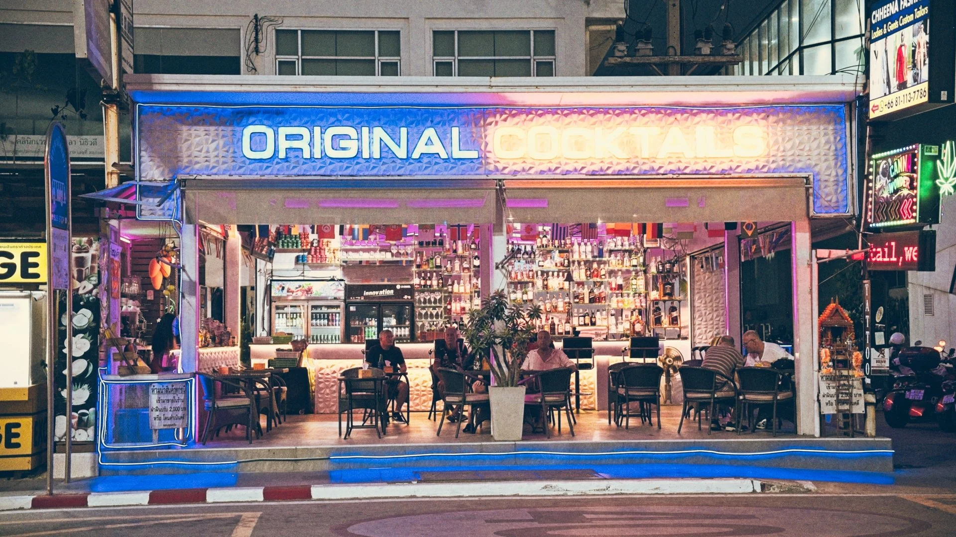 Nighttime view of a bar named 'Original Cocktails' with a neon-lit sign, outdoor seating, patrons sitting at tables, and a display of bottles inside the bar.